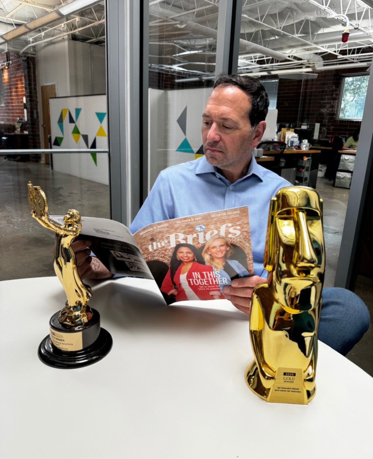 Man reading a magazine at a table with two gold trophies, one shaped like a person and the other like a face.