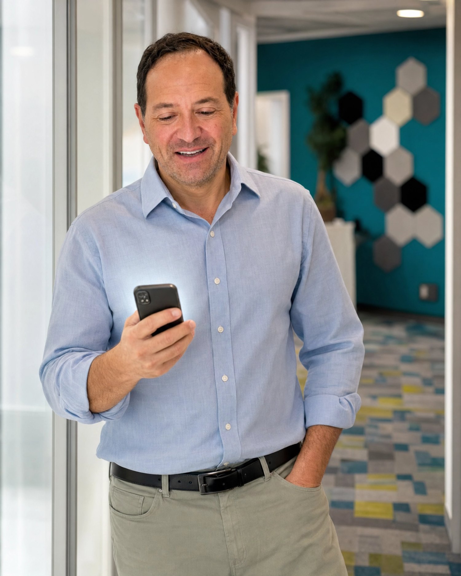 Man in light blue shirt looking at smartphone in modern office corridor, smiling, with geometric wall art in background.