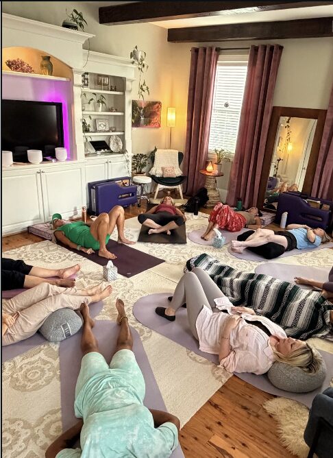 People lying on mats in a living room during a yoga class, with shelves, a mirror, and windows with curtains.