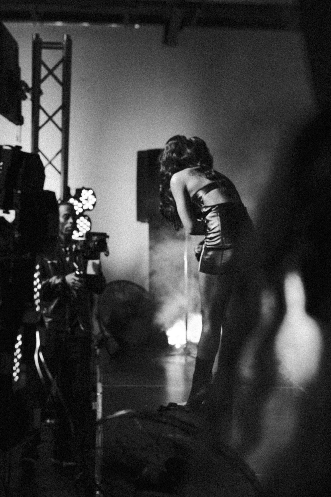 A woman in a dress stands on stage, with equipment and people around, in a black and white setting.