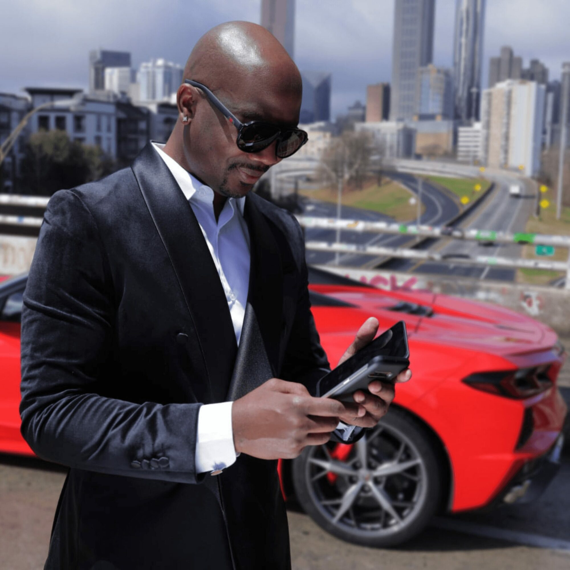 Man in sunglasses and suit using a smartphone outdoors with a red sports car and city skyline in background.