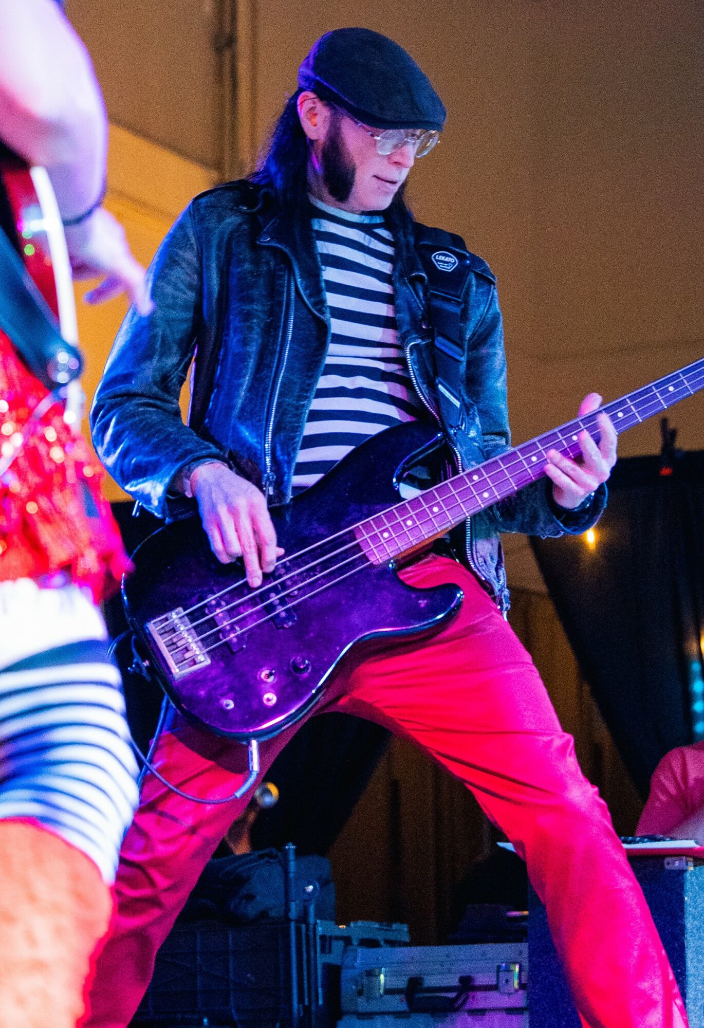 Person playing a purple bass guitar, wearing a black leather jacket, striped shirt, red pants, and a black cap.