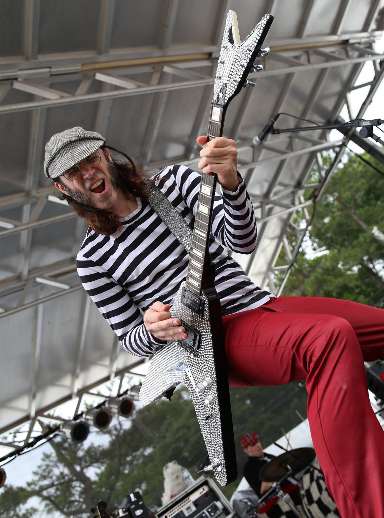 Person playing a large, pixelated guitar on stage, wearing a striped shirt, red pants, and a hat, smiling.