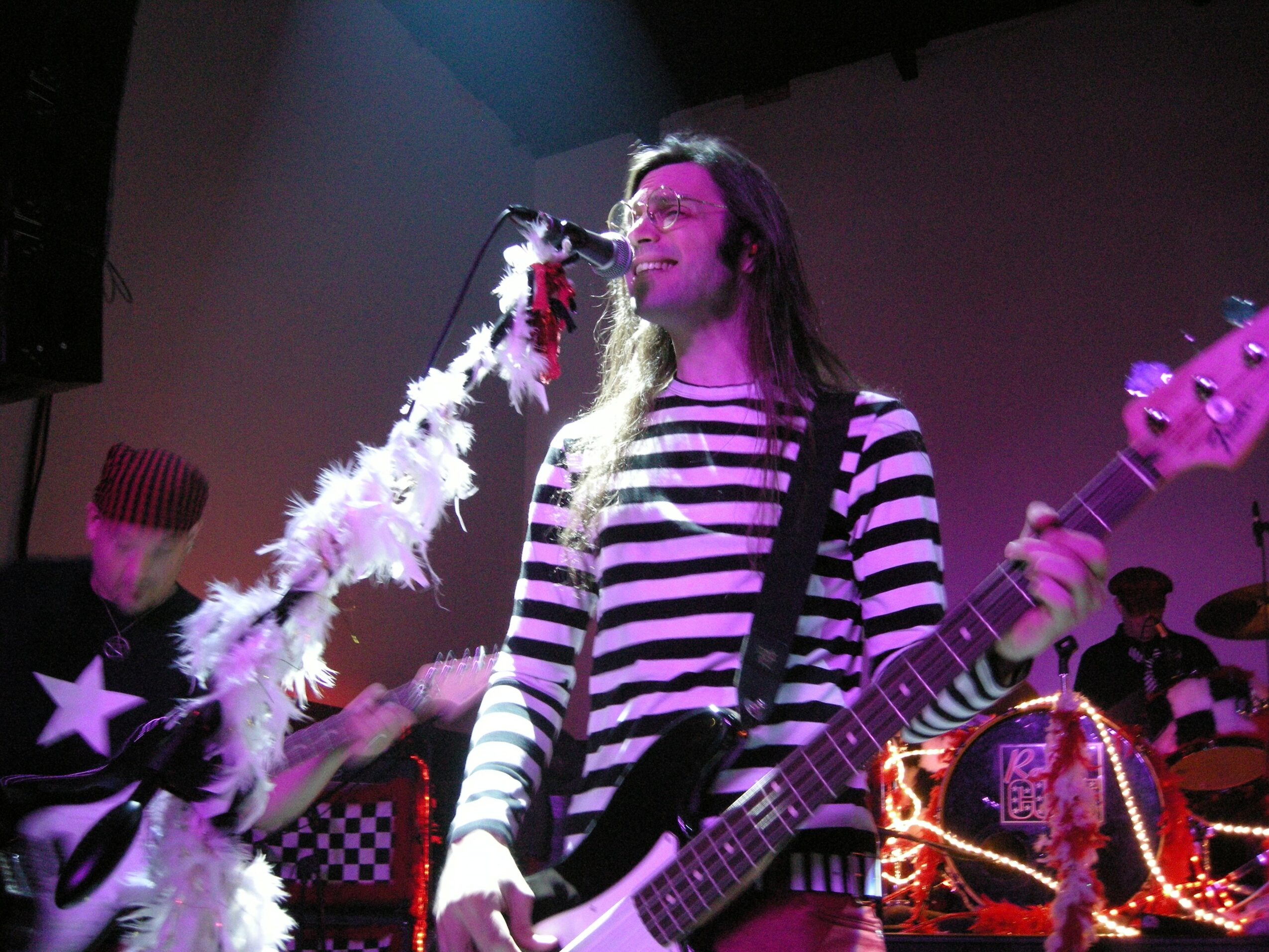 Person with long hair wearing a striped shirt playing guitar and singing on stage with colorful lights.