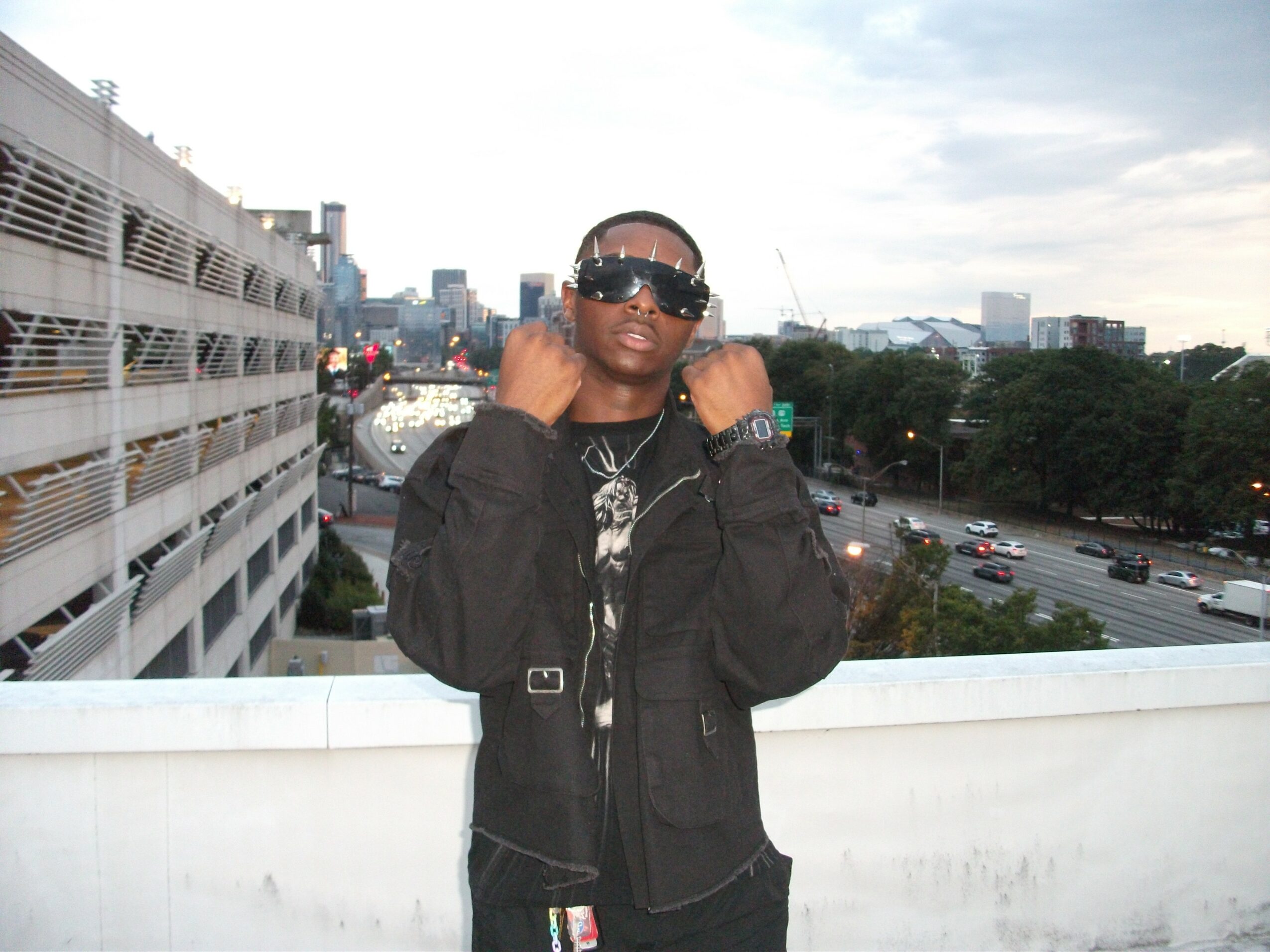 Person wearing sunglasses and a black jacket standing outdoors on a balcony with cityscape and traffic in the background.