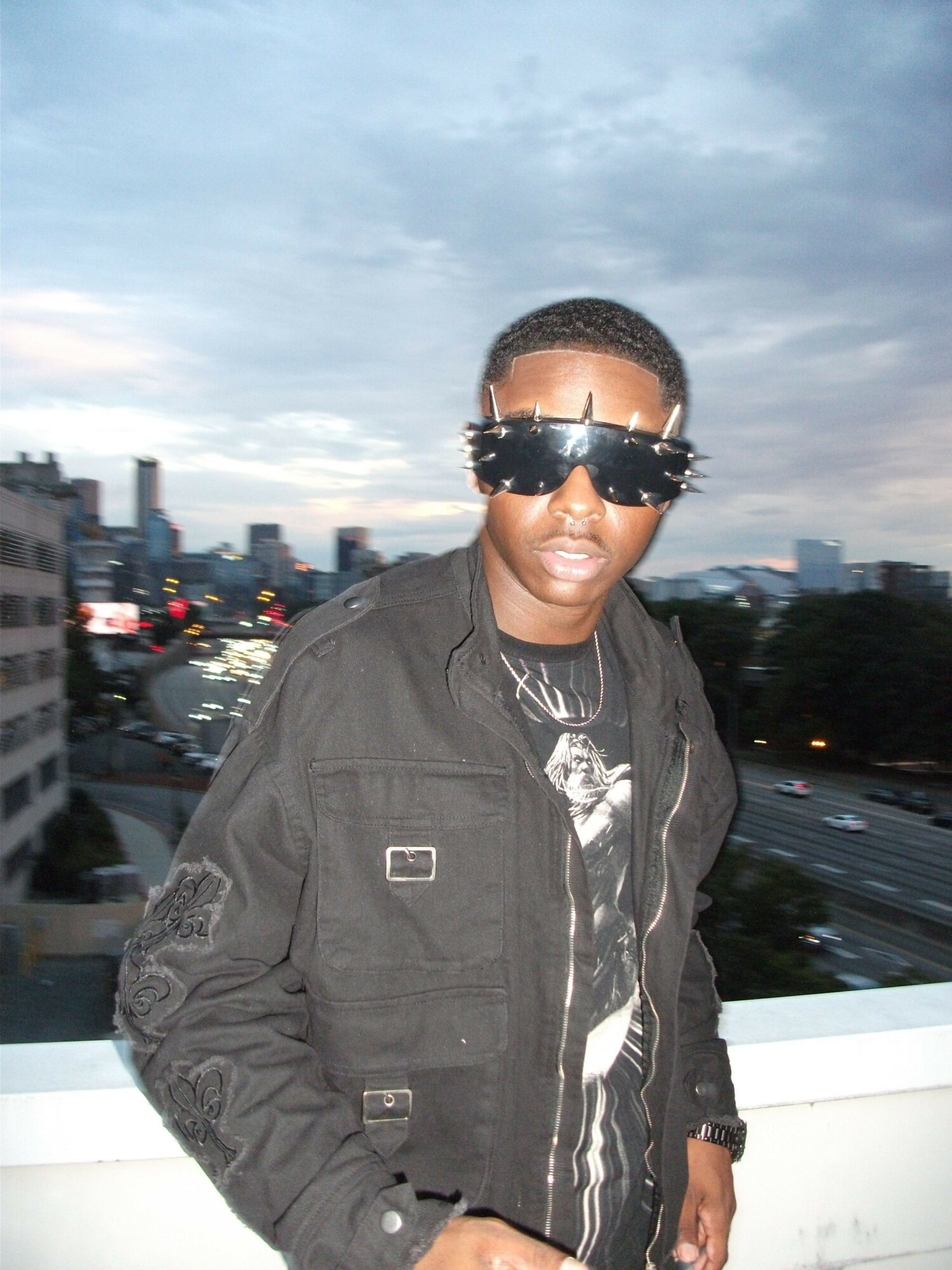 Person wearing sunglasses and a dark jacket standing outdoors with city buildings and sky in background.