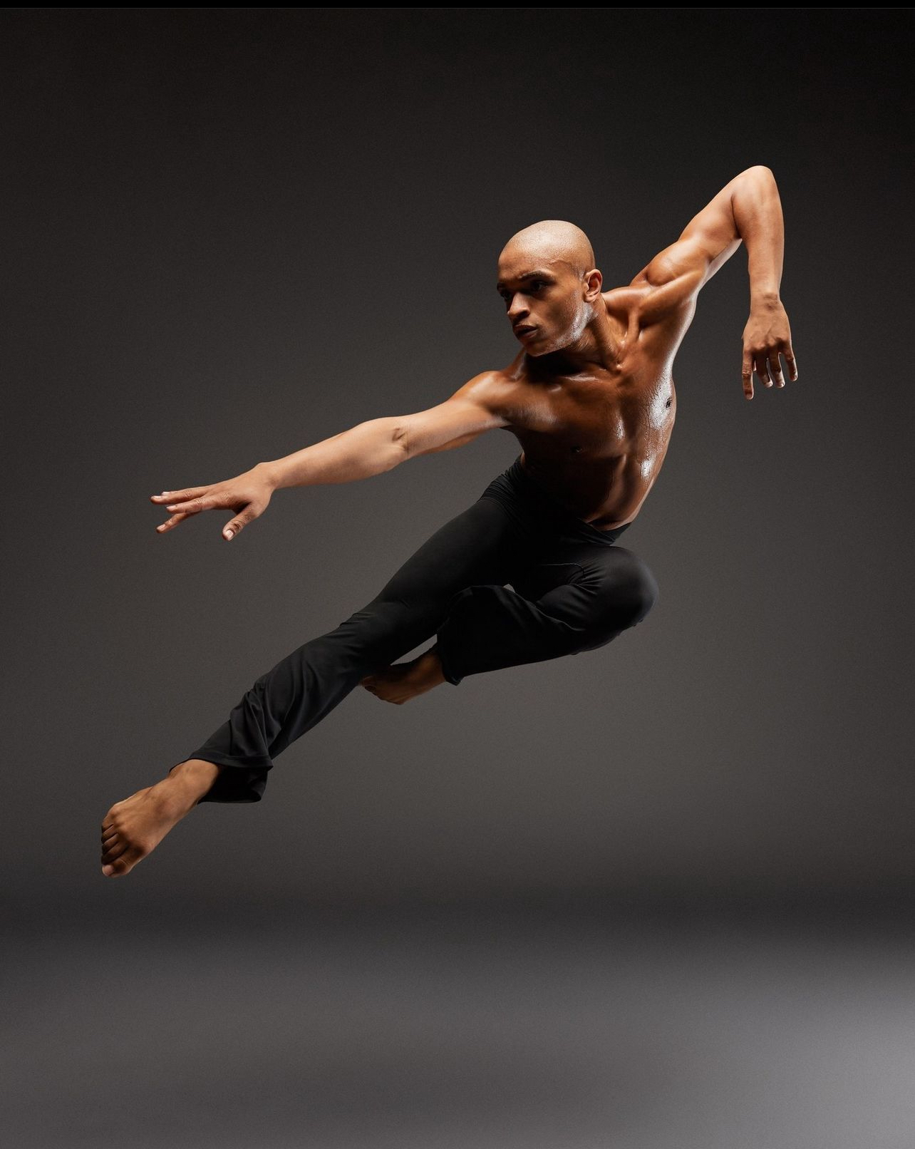 Bald male dancer in black pants leaps with arms extended, against a dark background.