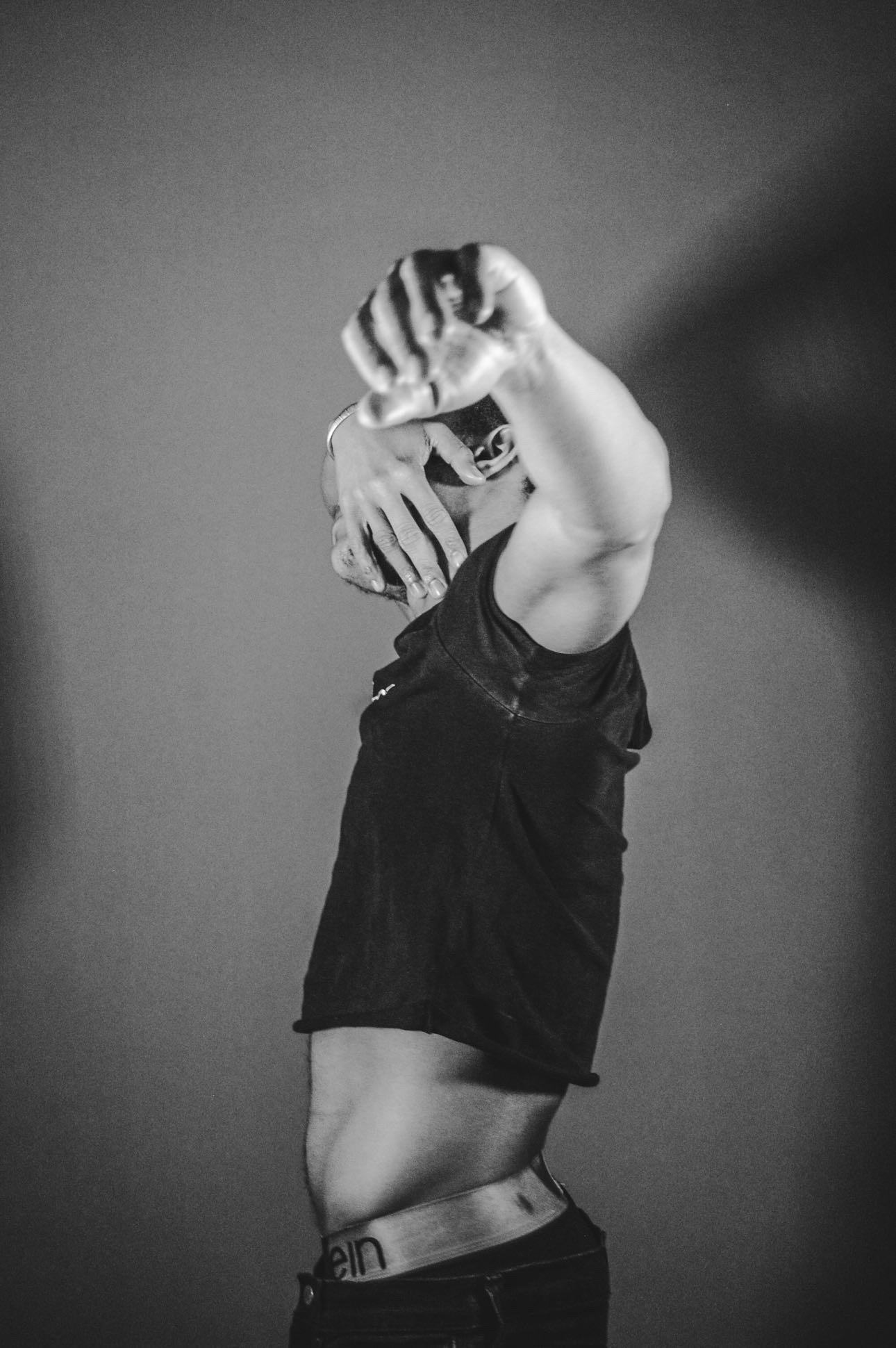 Person with arms raised, covering face, wearing a black shirt, in black and white, against a plain background.
