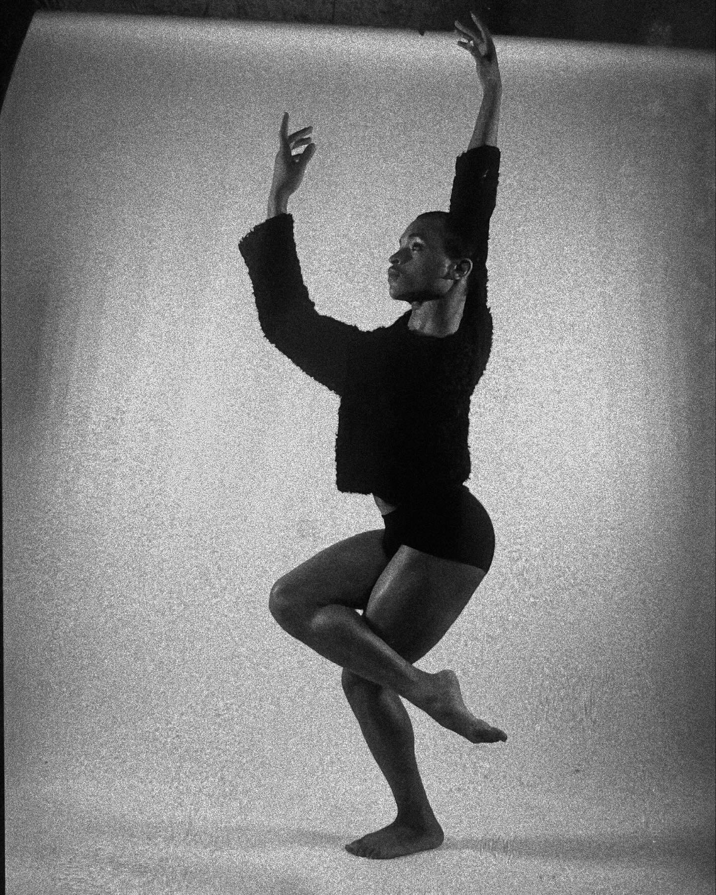 Person performing a dance pose with arms raised and one leg bent, against a plain background.