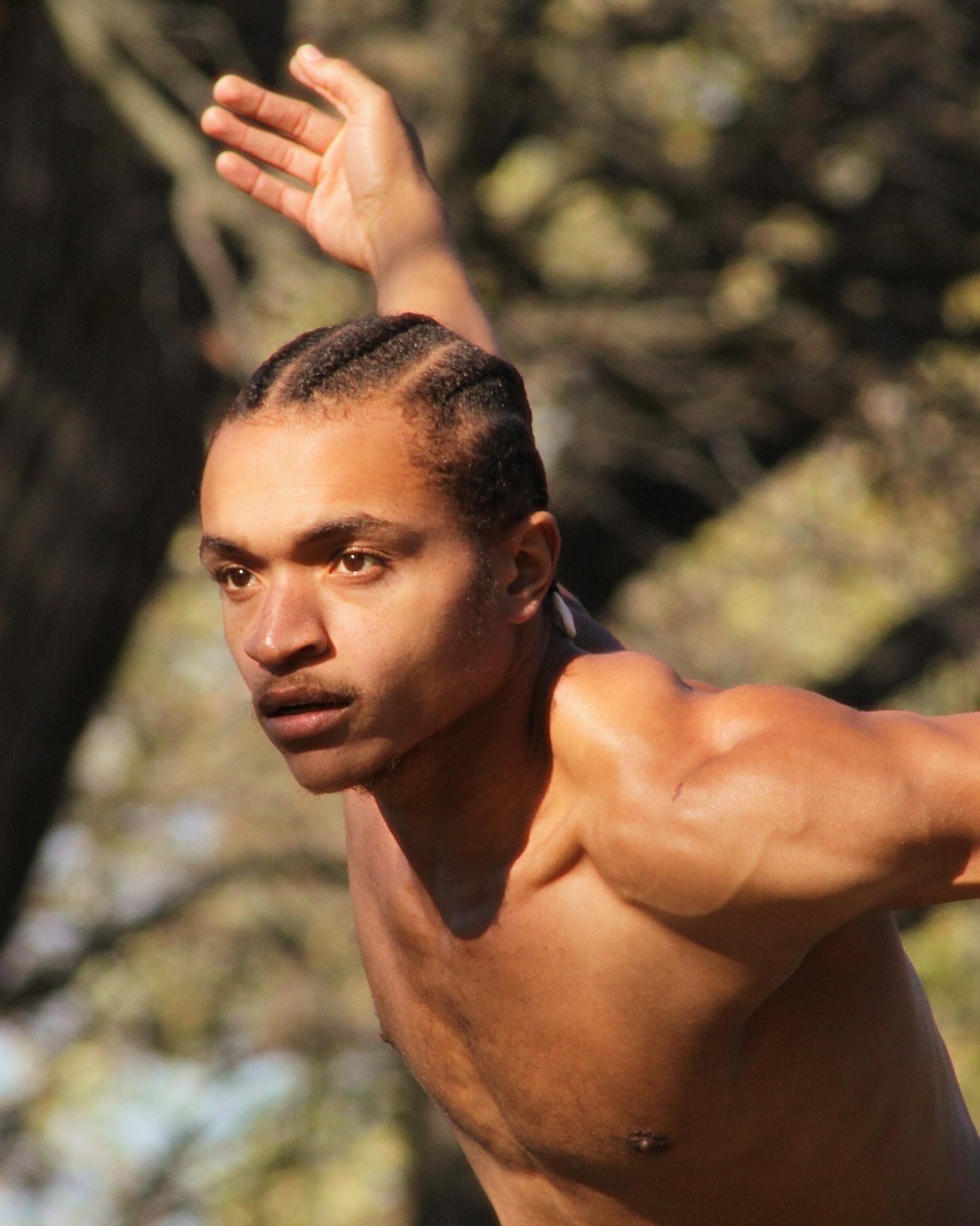 A shirtless man with braided hair extends his arm forward outdoors with trees in the background.