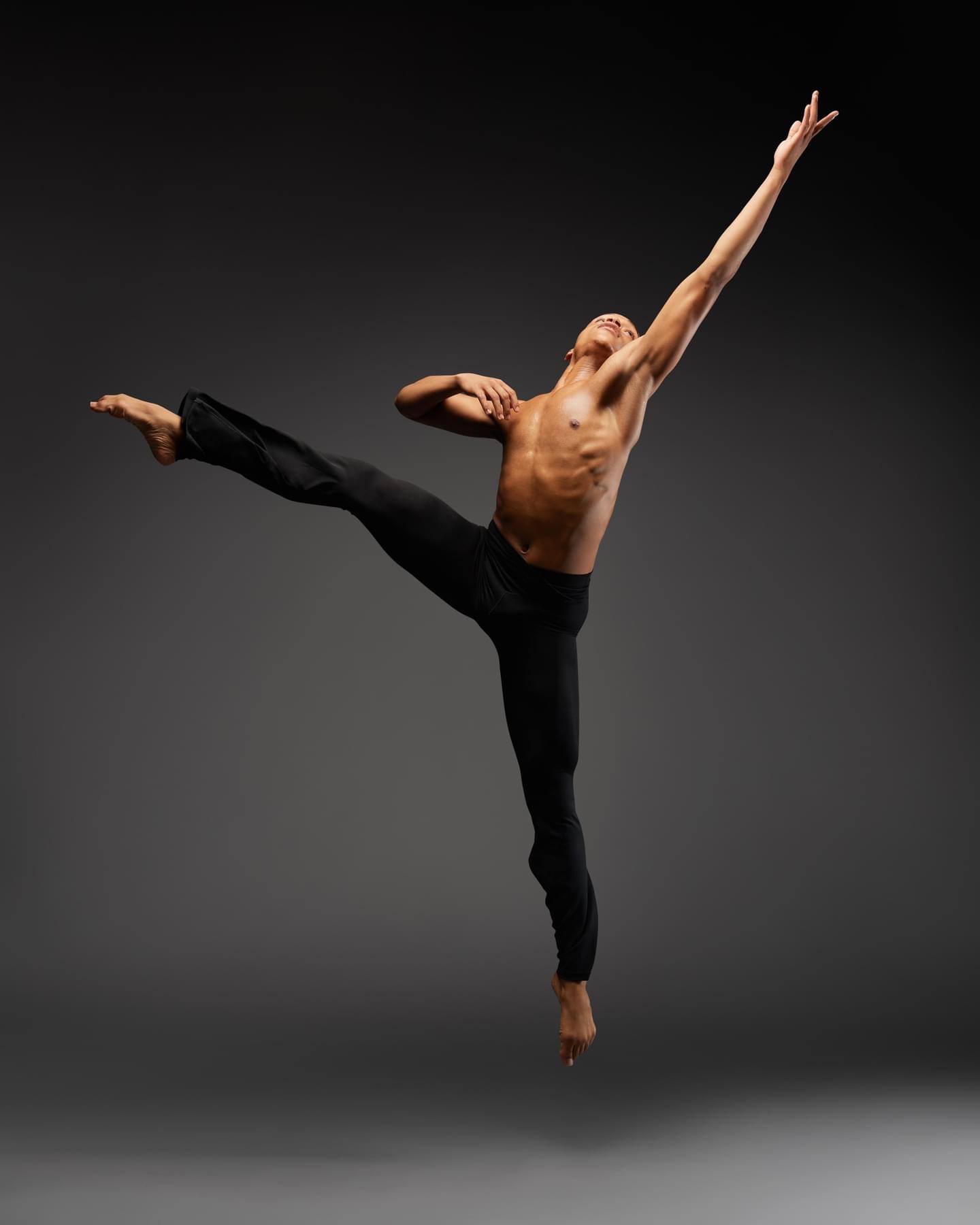 A male dancer performs a high leap with one arm extended upward and the other bent, against a dark background.