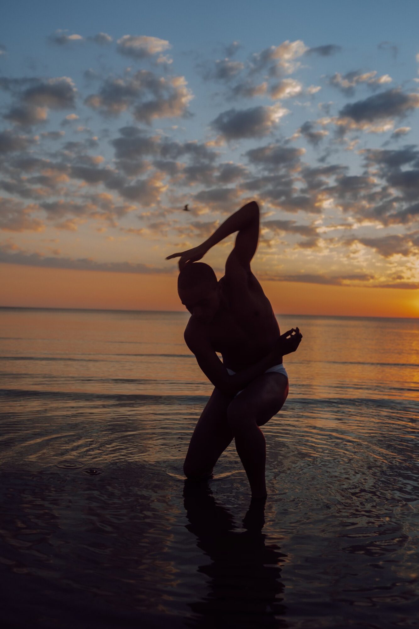 Person standing in water at sunset, silhouette against sky with clouds, holding a pose with one leg raised.