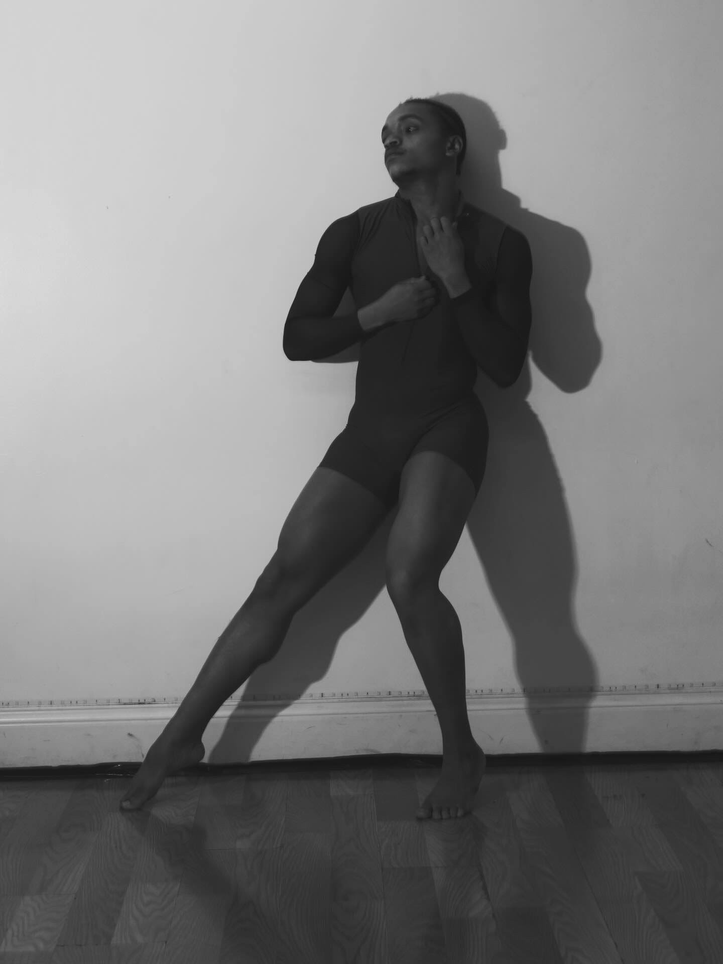 Person standing against wall, one leg bent, hands near face, shadow cast on wall, in black and white.