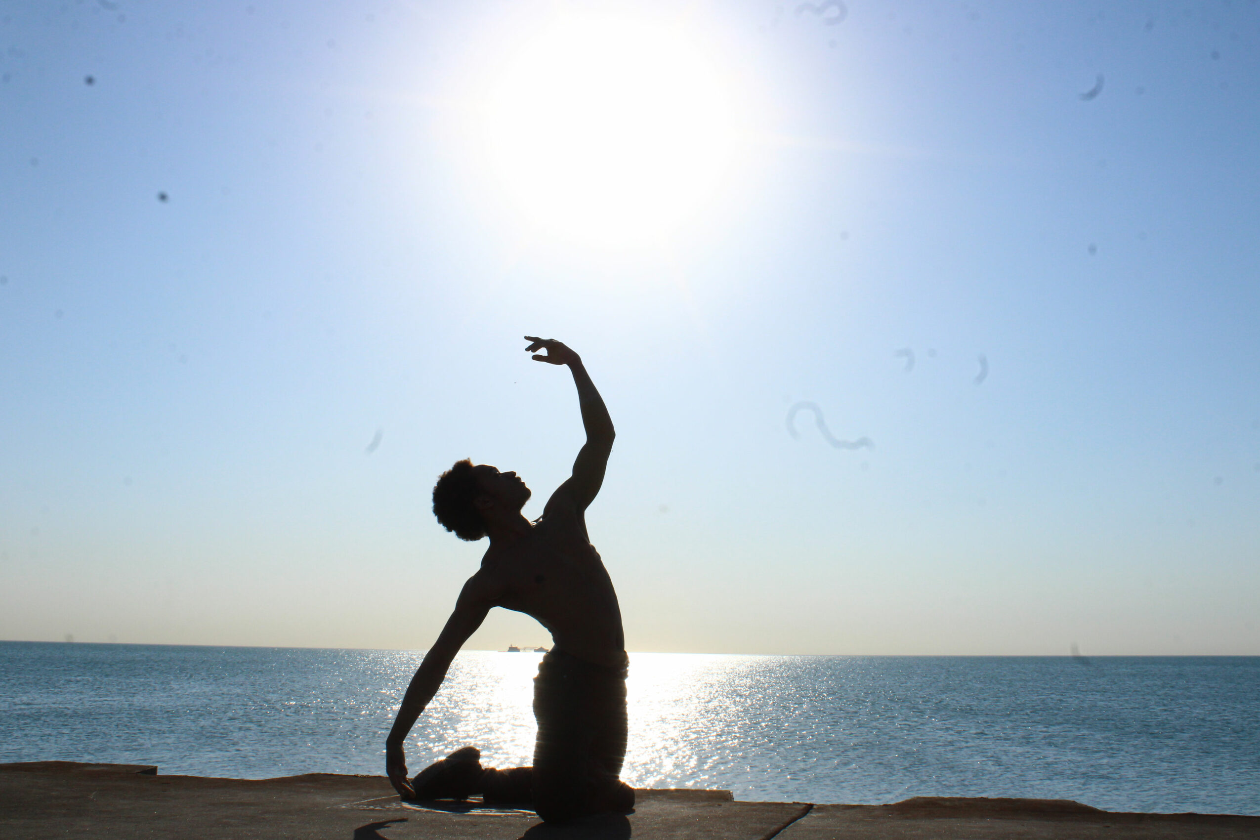 Silhouette of a person kneeling on the beach with one arm raised, facing the sun over the ocean.
