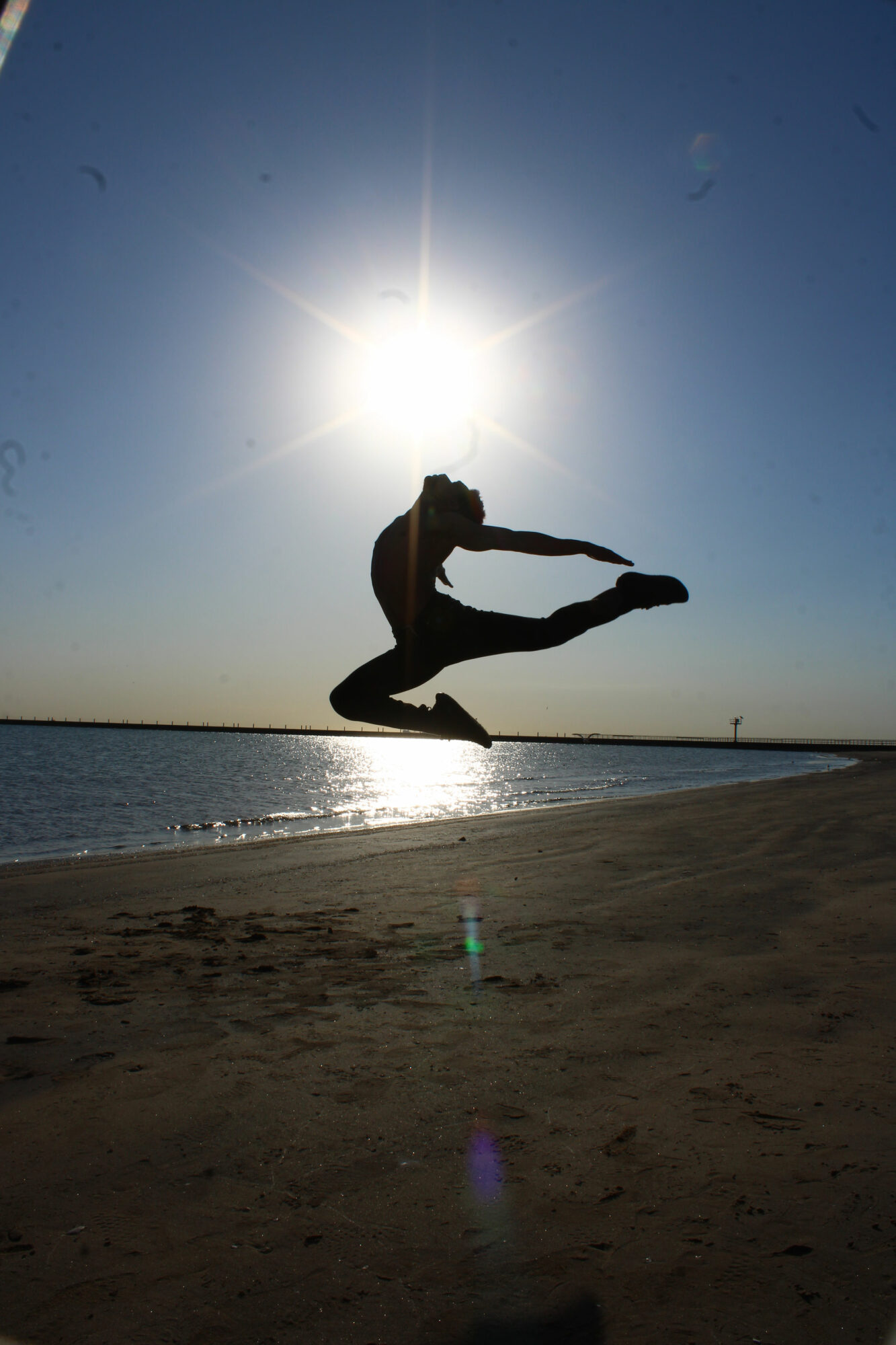 Person jumping on beach with the sun behind, creating a silhouette and lens flare, over water and sky.