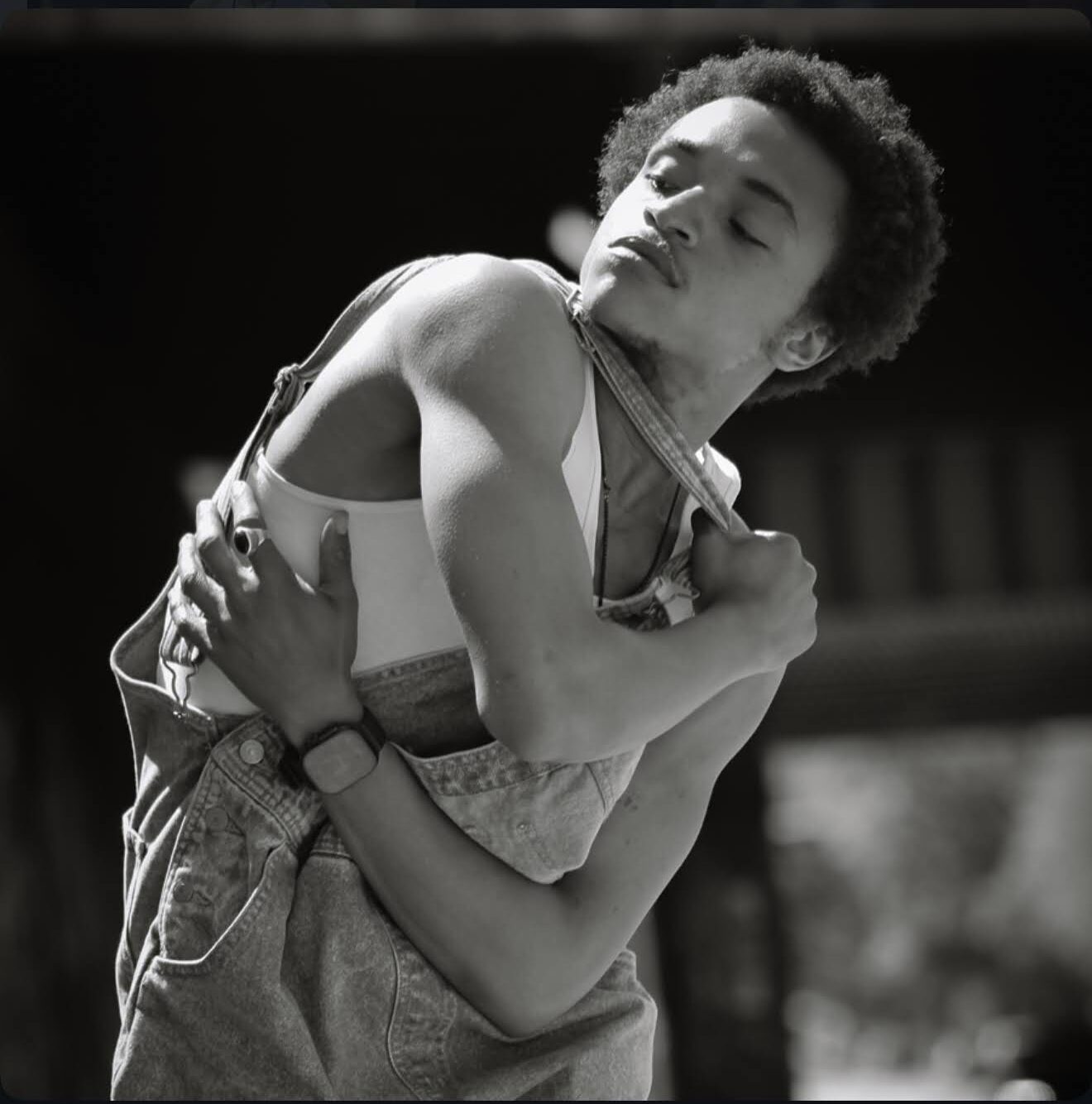 Young person with curly hair adjusting clothing, eyes closed, outdoors, black and white photo.