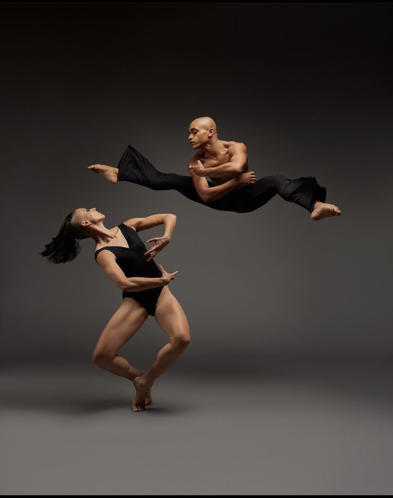 Two dancers perform mid-air against a dark background, one balancing on one leg and the other leaping horizontally.