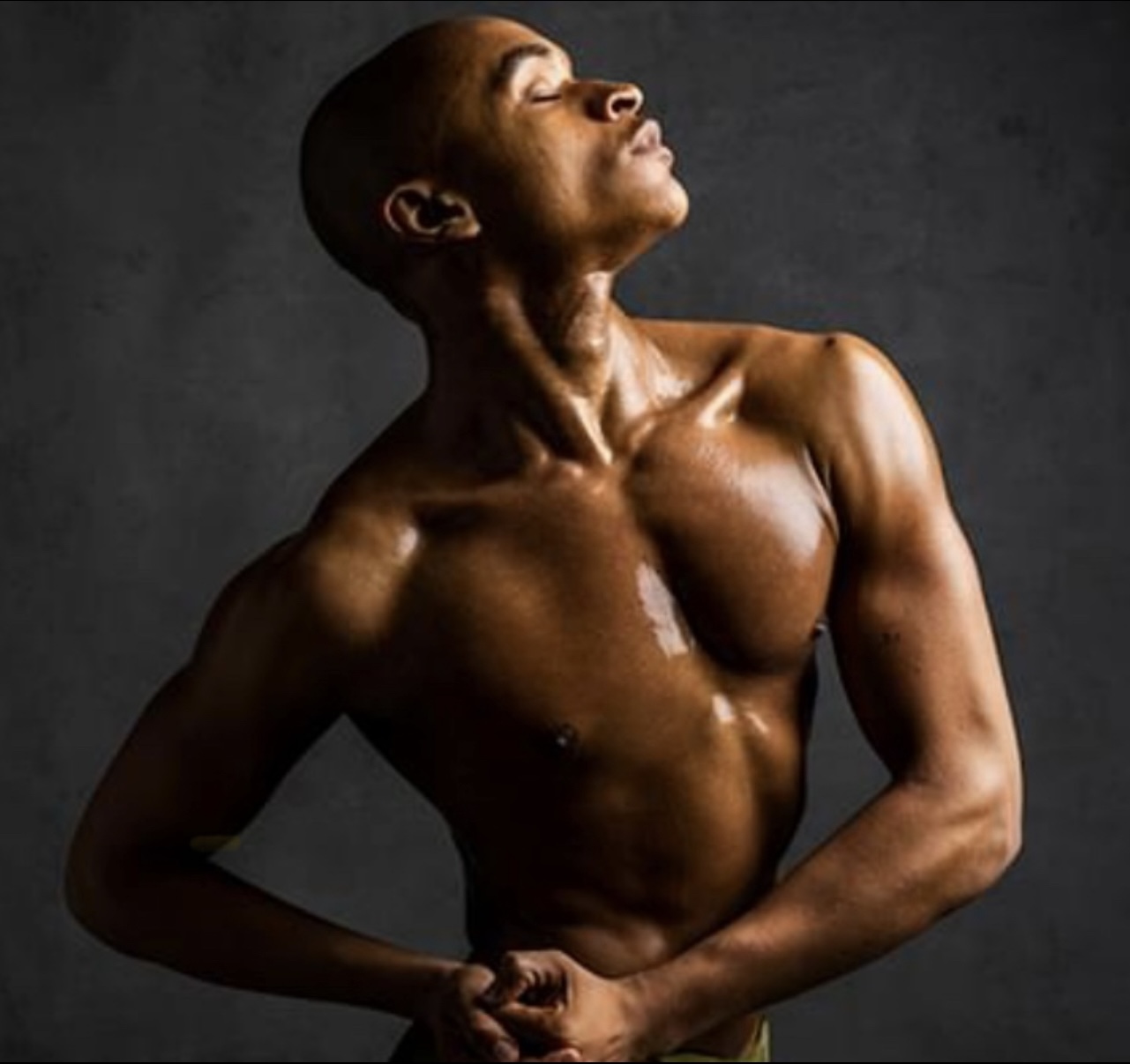 A shirtless person with a muscular build poses with head tilted back and eyes closed against a dark background.