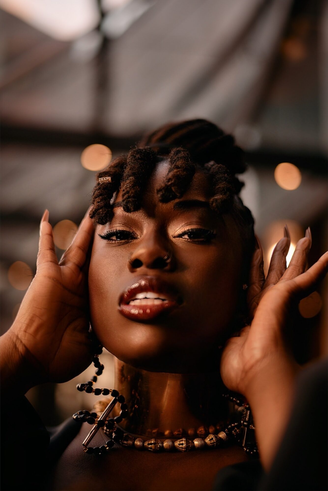Close-up of a woman with styled hair and jewelry, holding her hands near her face, in a dimly lit setting.
