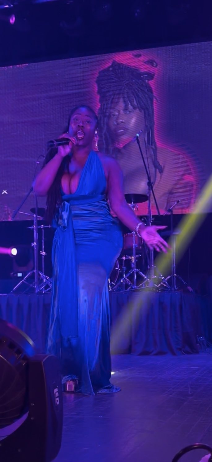 A woman singing on stage with a large screen behind her showing her image, wearing a blue dress, with a yellow spotlight.