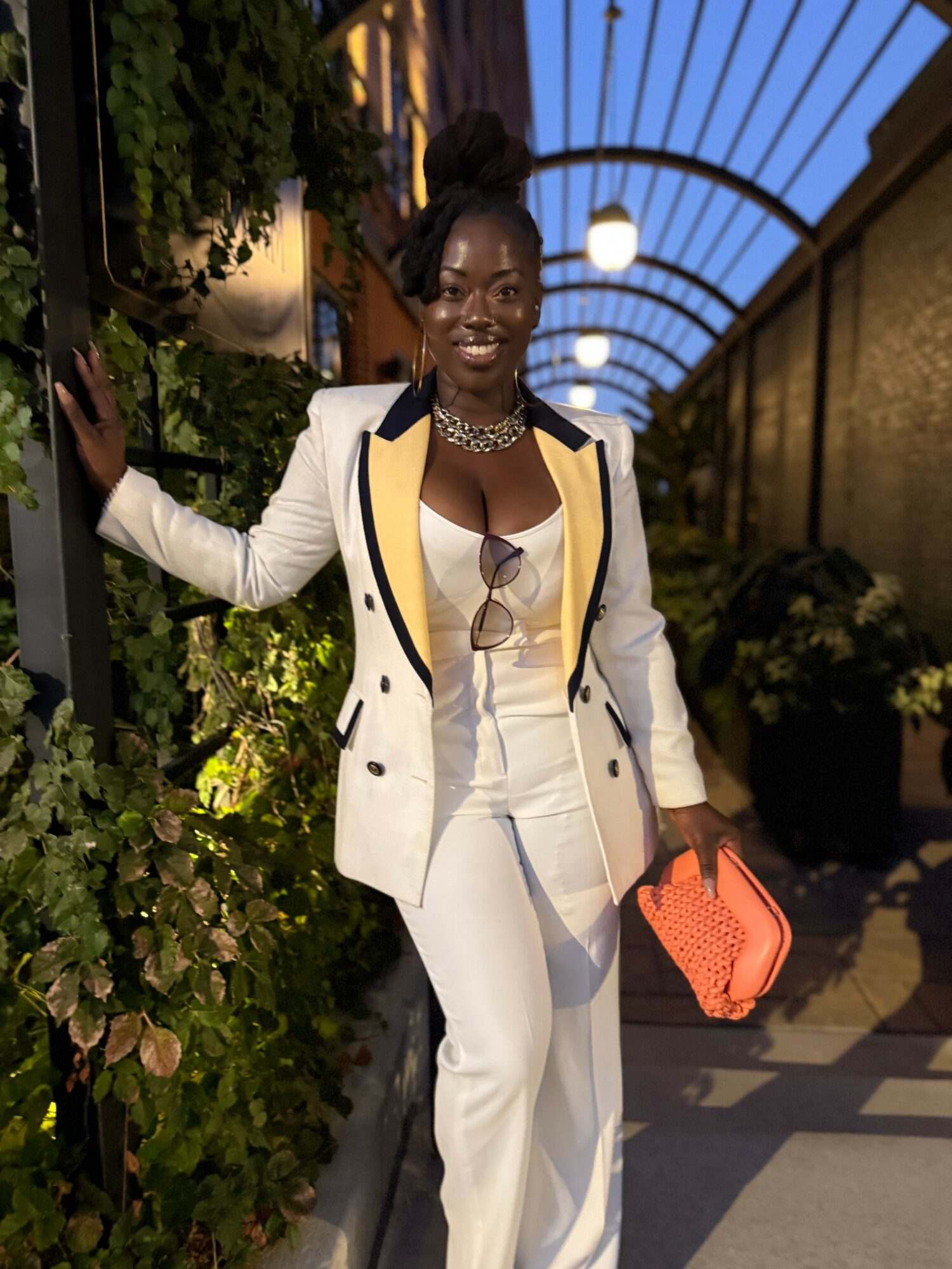 Woman in white suit with yellow accents, holding a coral bag, standing outdoors under a curved glass roof, smiling.