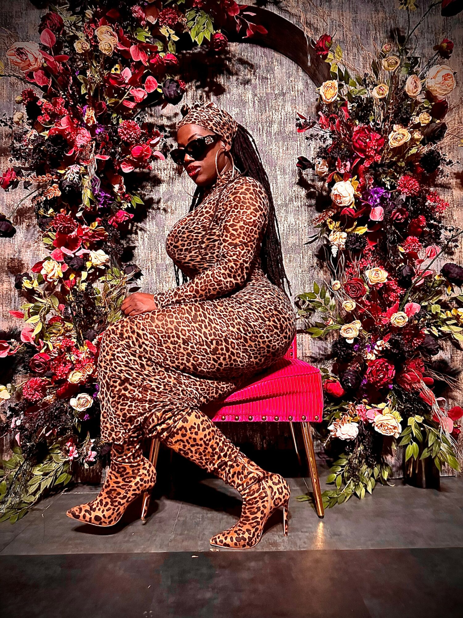 Woman in leopard print dress and heels sitting on a pink chair surrounded by colorful flowers, wearing sunglasses.