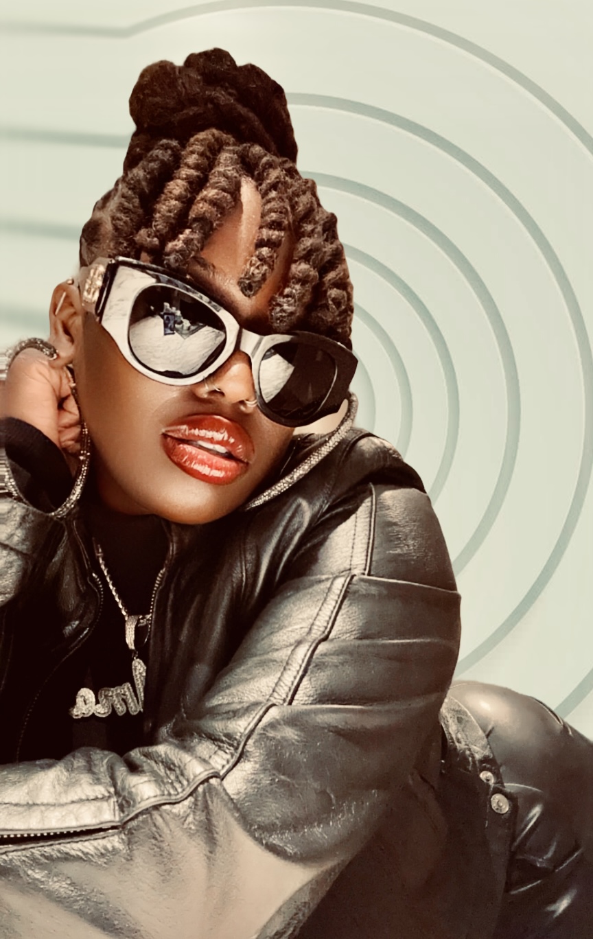 Woman with styled hair, large sunglasses, red lipstick, wearing a leather jacket, smiling, against a light background.