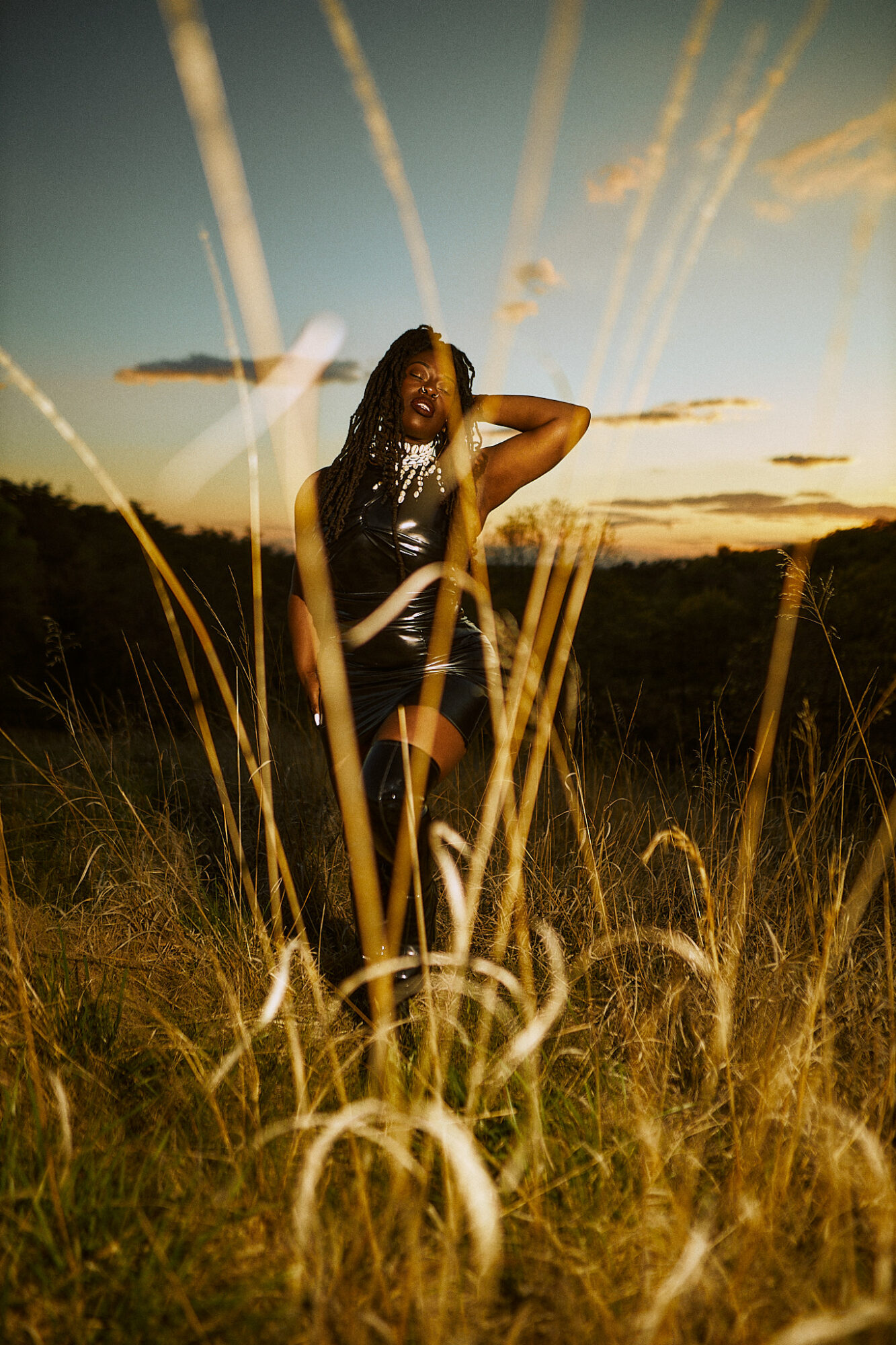 Person standing in a grassy field during sunset, with tall grass in the foreground and a clear sky with streaks of clouds.