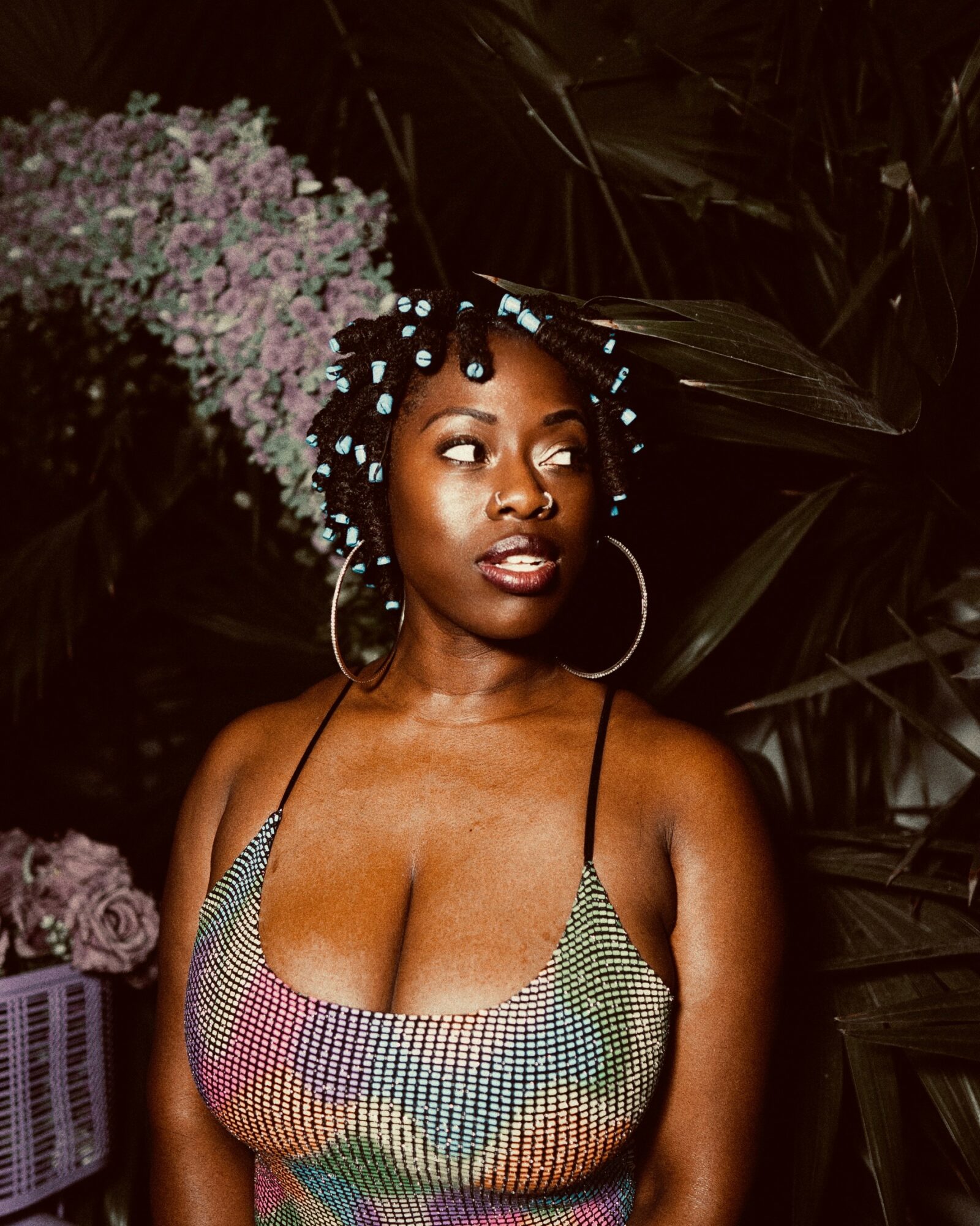 Woman with curly hair and hoop earrings in a colorful top, surrounded by dark foliage and purple flowers.