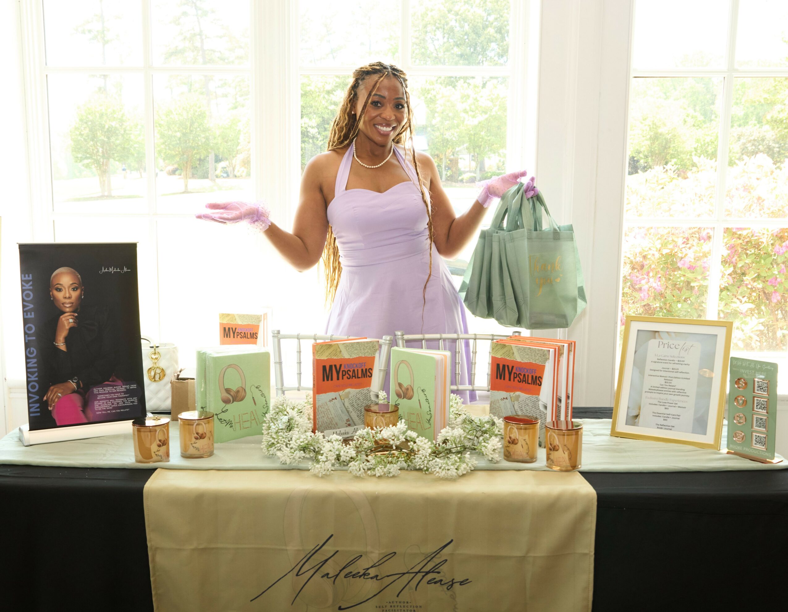 Woman standing behind a table with books and framed photos, smiling, in a bright room with large windows.