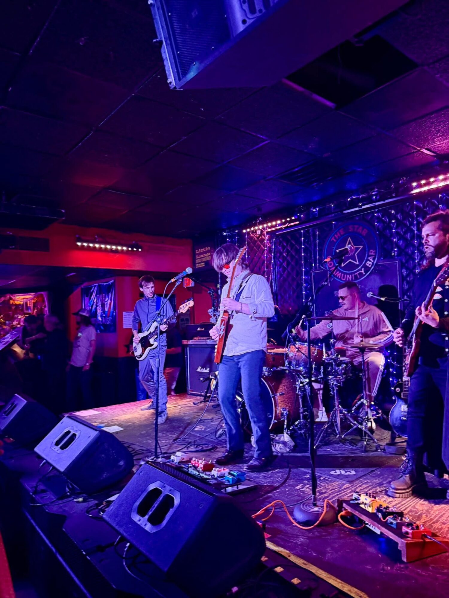 Band performing live on stage with instruments and colorful lighting, audience visible in the background.