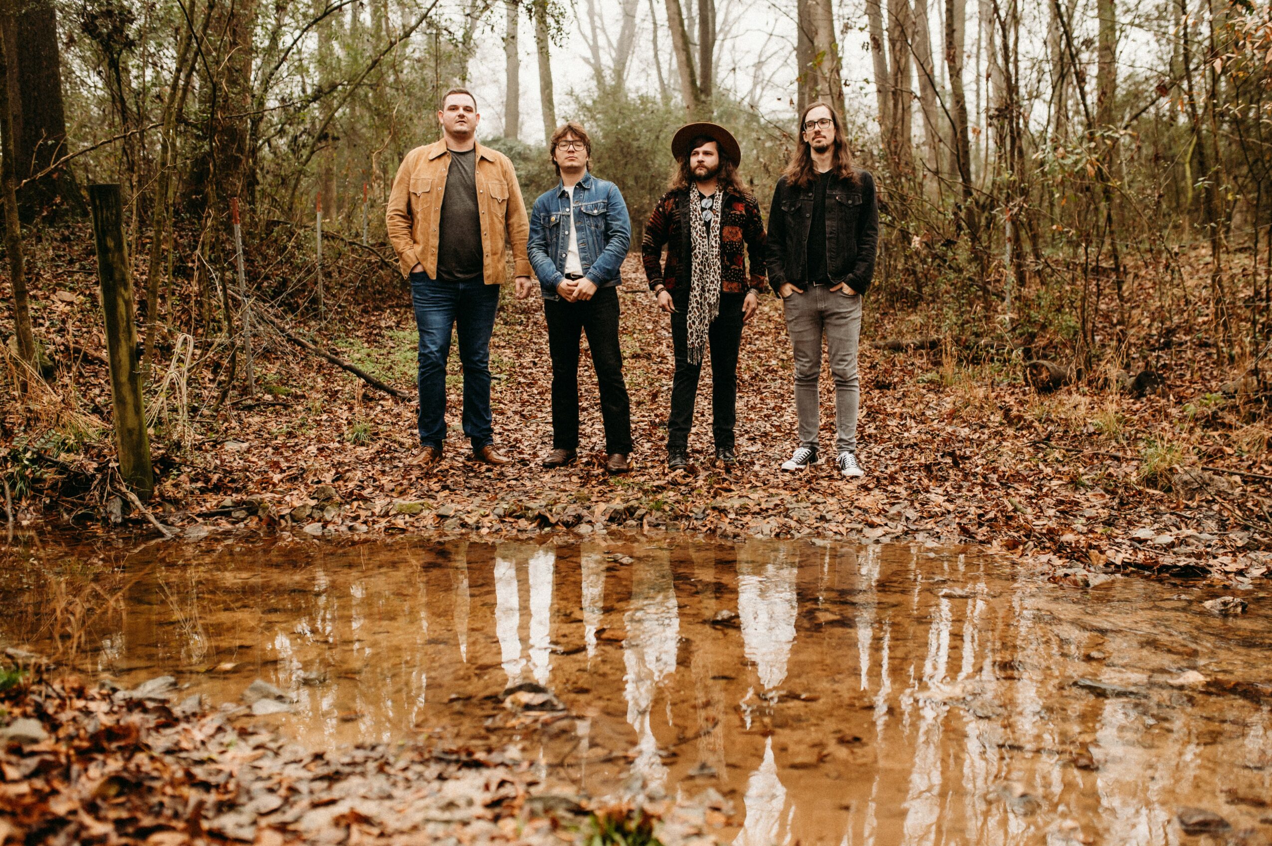Four people stand in a forest near a reflective body of water, with trees and fallen leaves in the background.