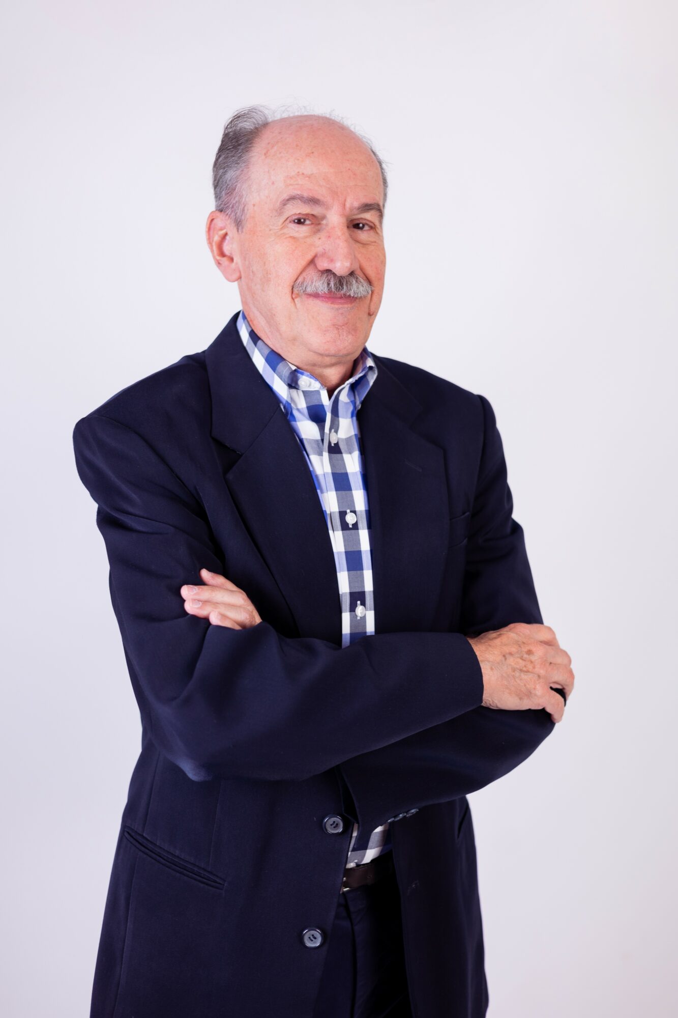 Older man with gray hair and mustache wearing a dark blazer and checkered shirt, crossing arms, smiling, against a plain white background.