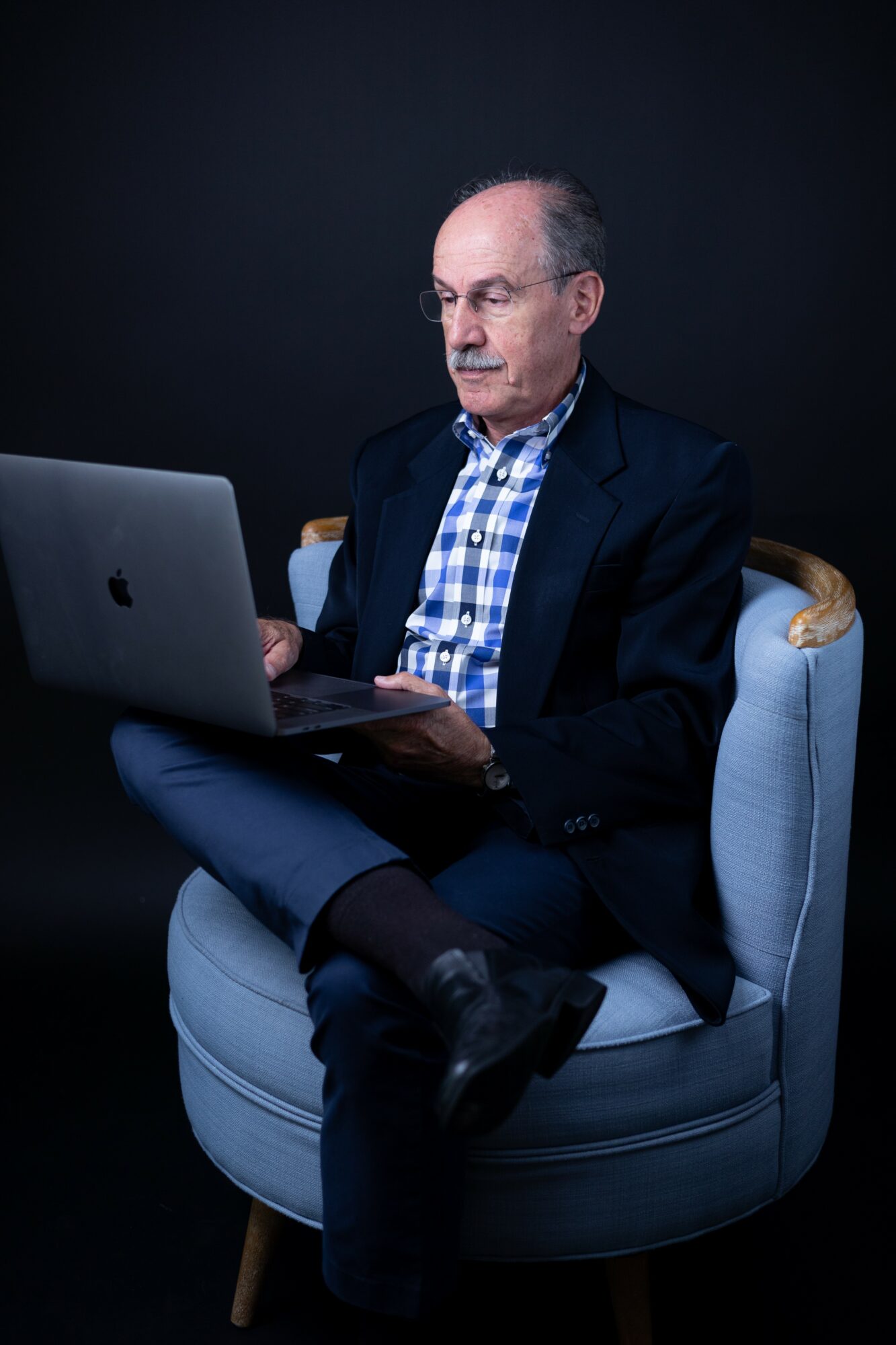 Older man with glasses in a dark suit and checkered shirt sitting on a light-colored chair, working on a laptop.