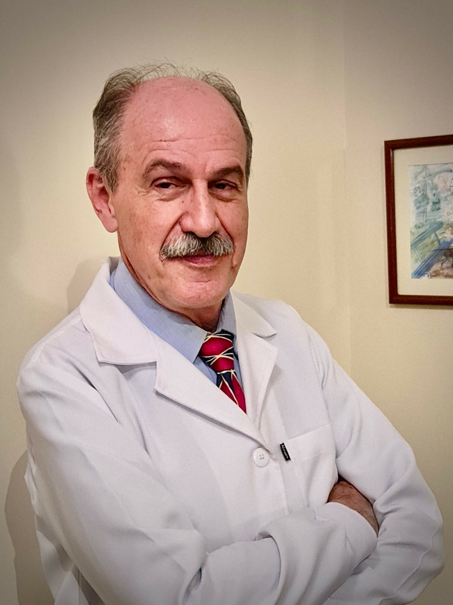 A middle-aged man with a mustache wearing a white lab coat and a striped tie, standing with arms crossed in a room with a framed picture on the wall.