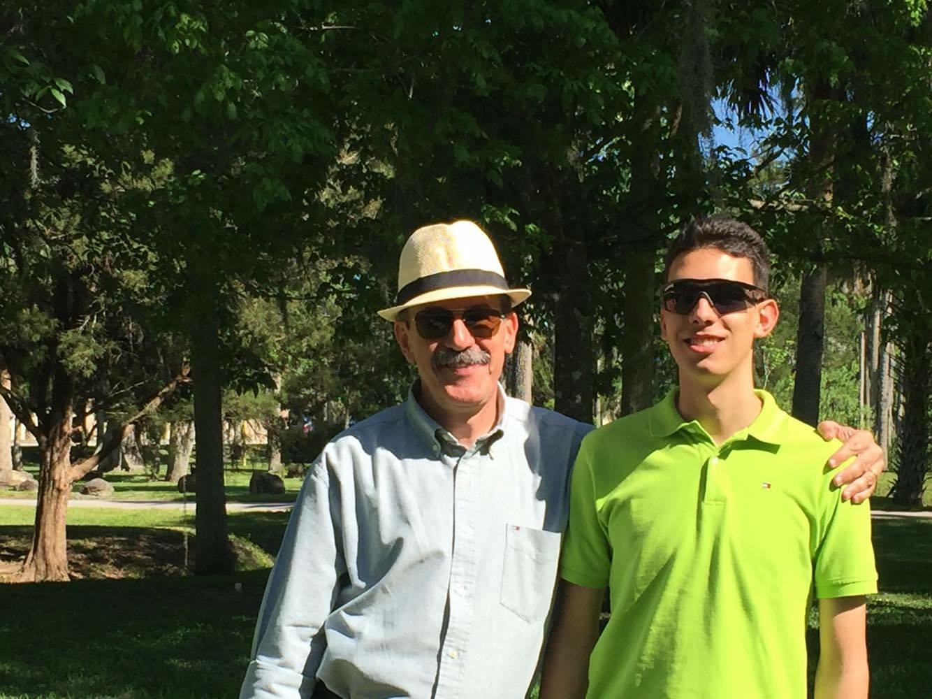Two men standing outdoors, one older with a hat and sunglasses, the other younger in a bright green shirt and sunglasses, smiling.
