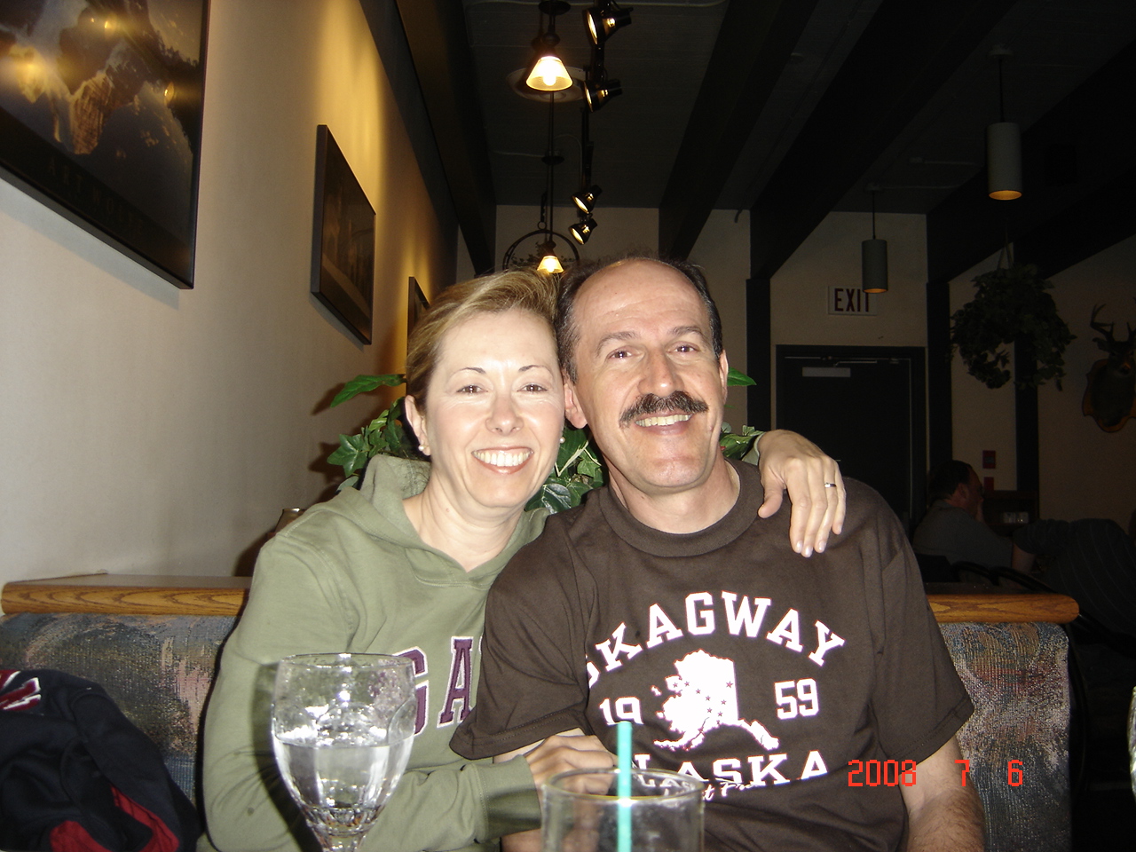 Two people smiling, one with arm around the other, sitting at a table with a glass of water, in a cozy indoor setting.