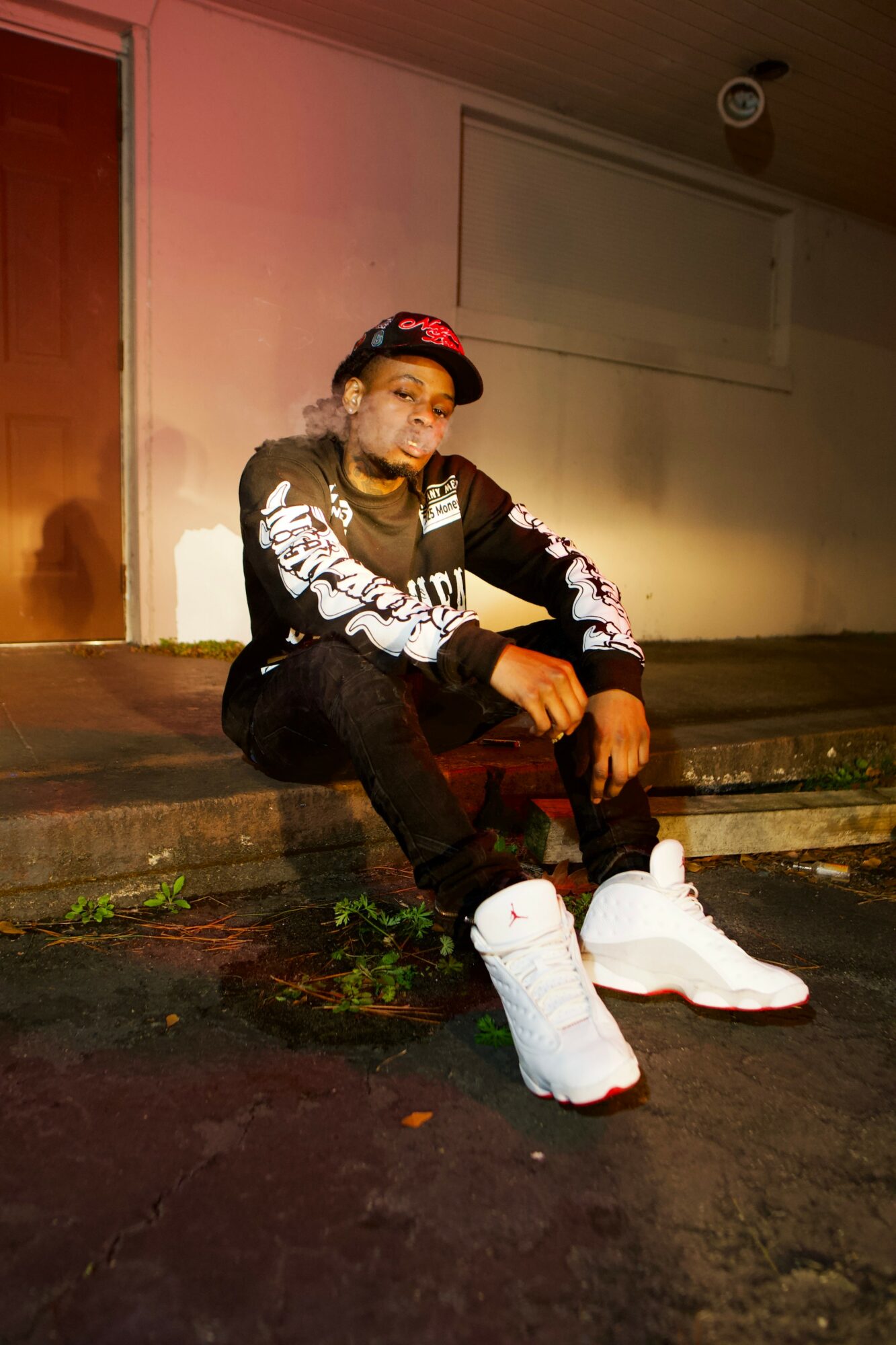 Young person sitting on a step outdoors at night, wearing a black and white jacket, black pants, and white boots.