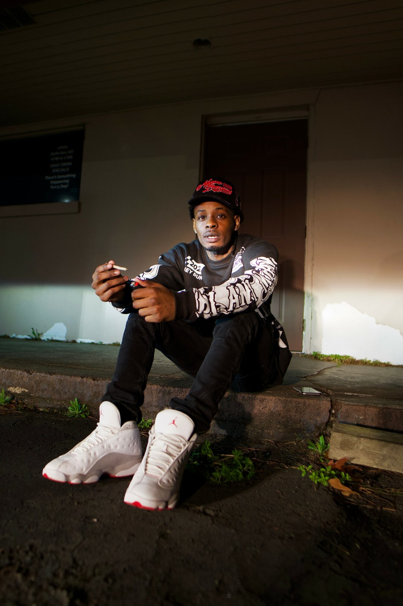 Young person sitting outdoors at night, wearing a red cap, black jacket, white sneakers, holding a phone, with a building in background.