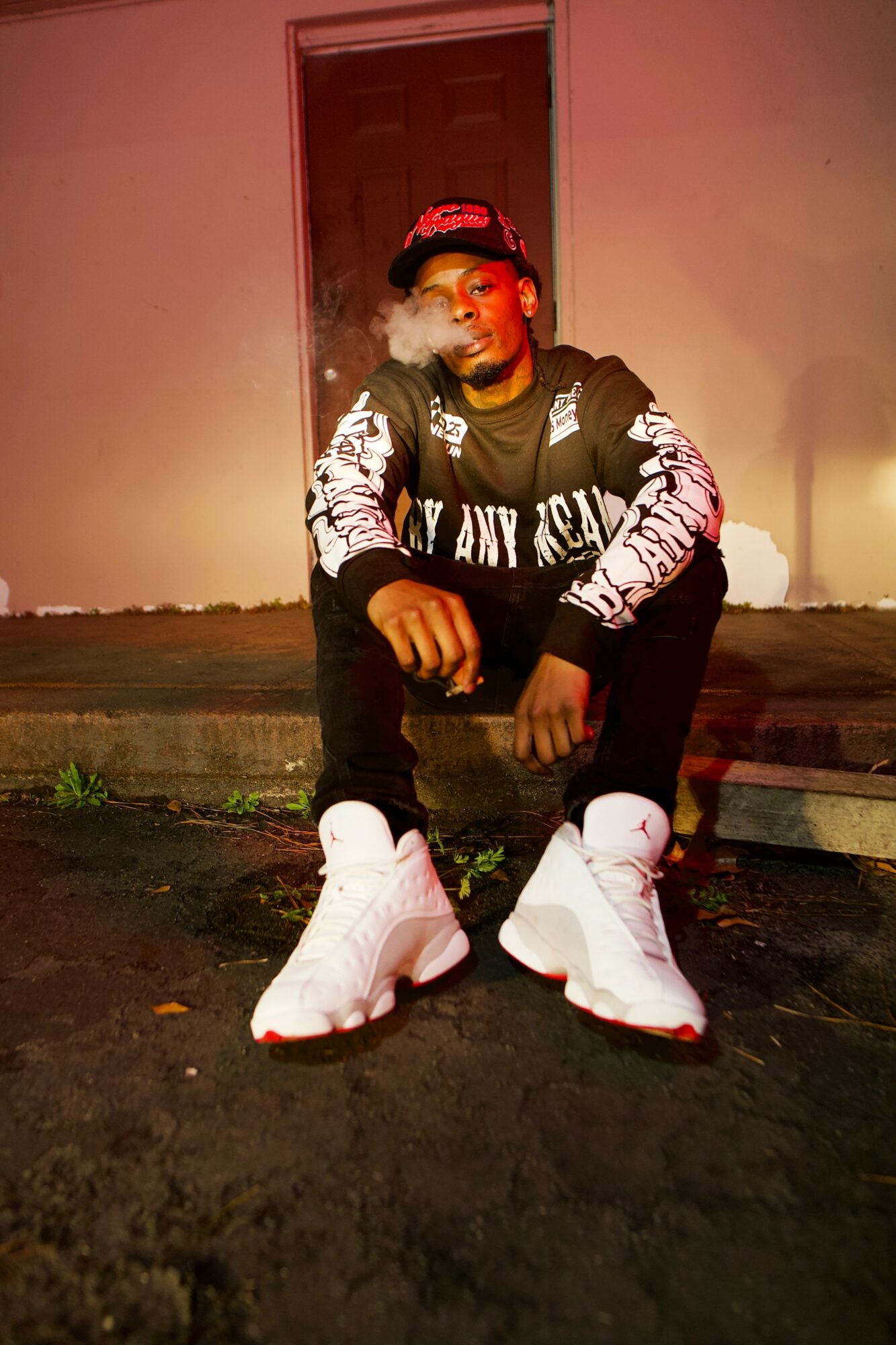 Person sitting on steps in front of a pink wall, wearing a black jacket, white sneakers, and a red cap, smoking.