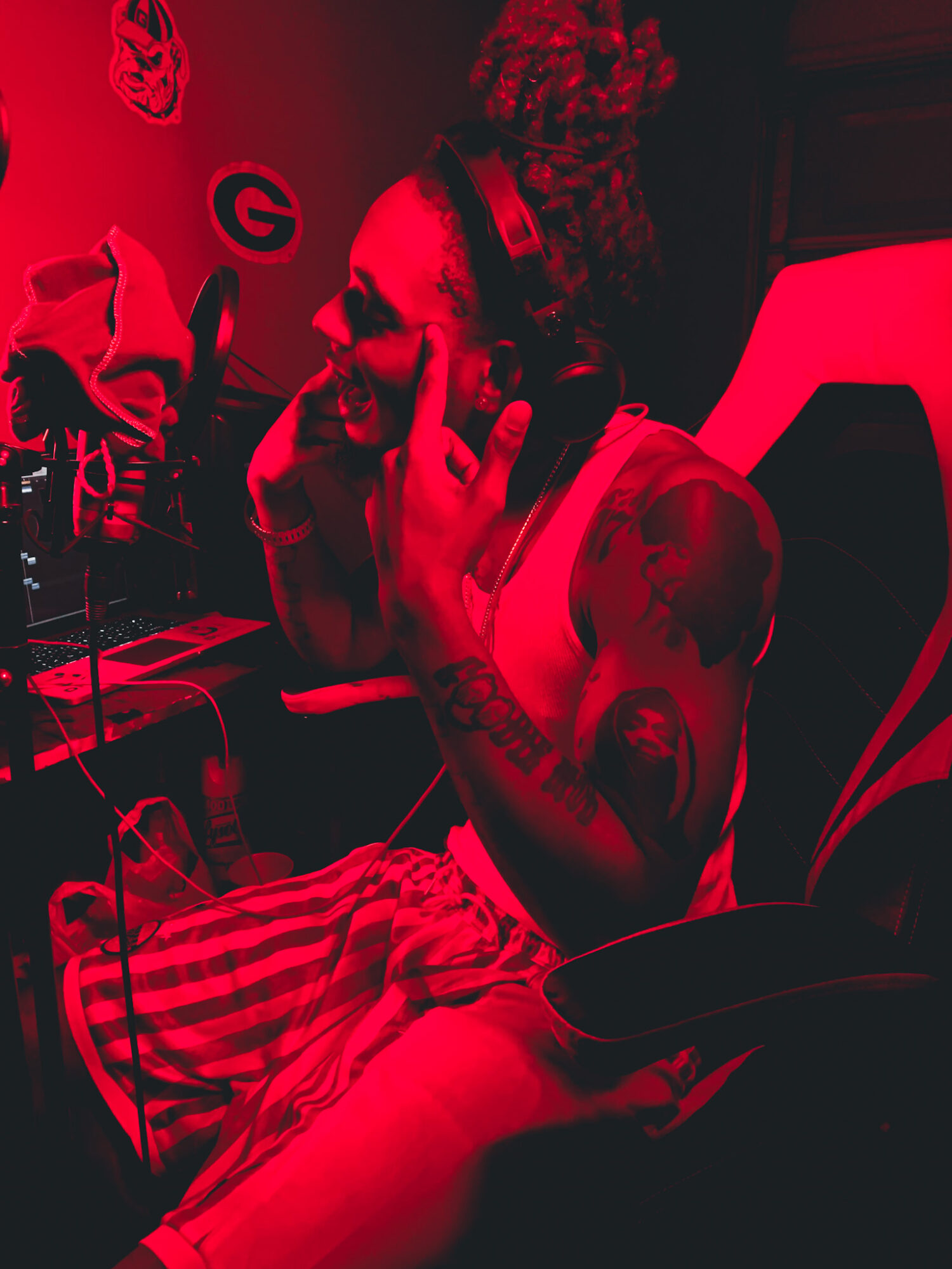 Person with tattoos and braided hair, sitting and talking on a phone, in a dimly lit environment with red lighting.