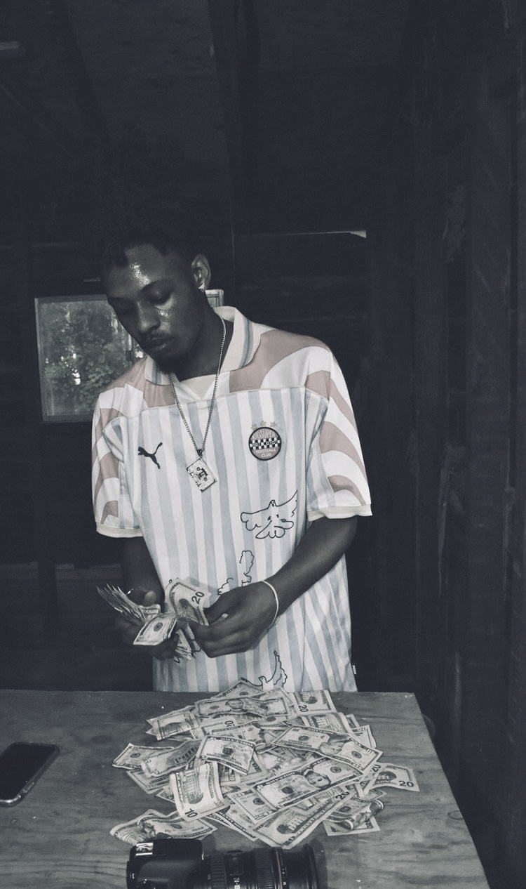 Young man in striped shirt stands behind a table with a large pile of cash, inside a dark room.