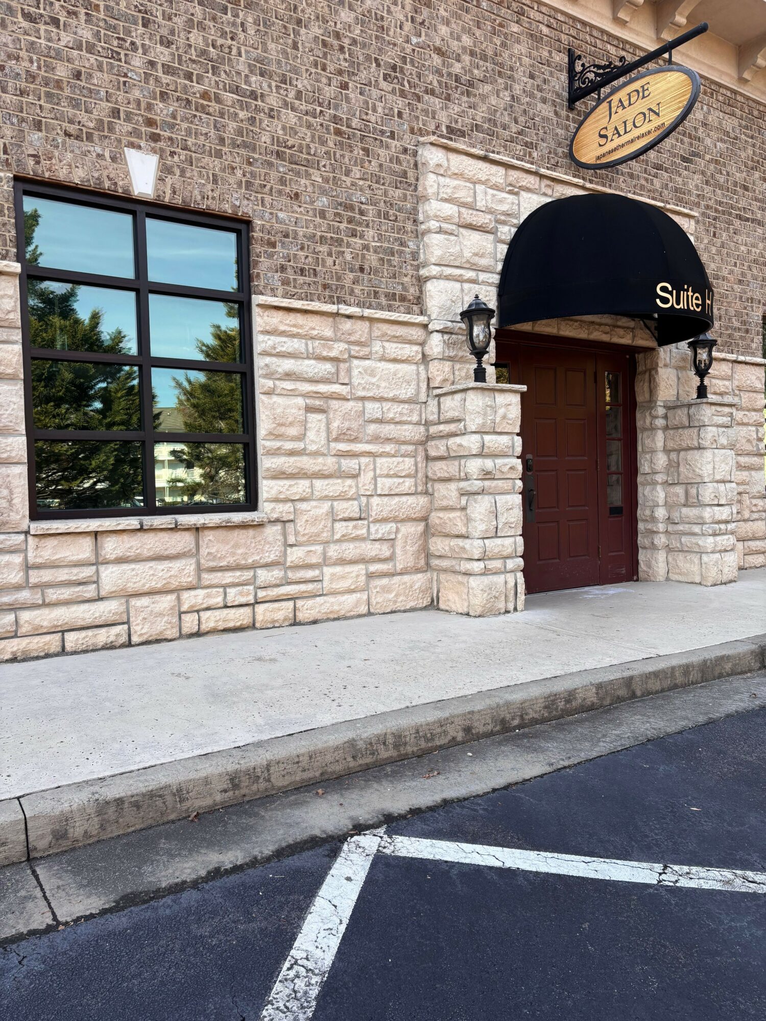 Building with brick and stone exterior, window reflecting trees, black awning over door, sidewalk, and parking lot.