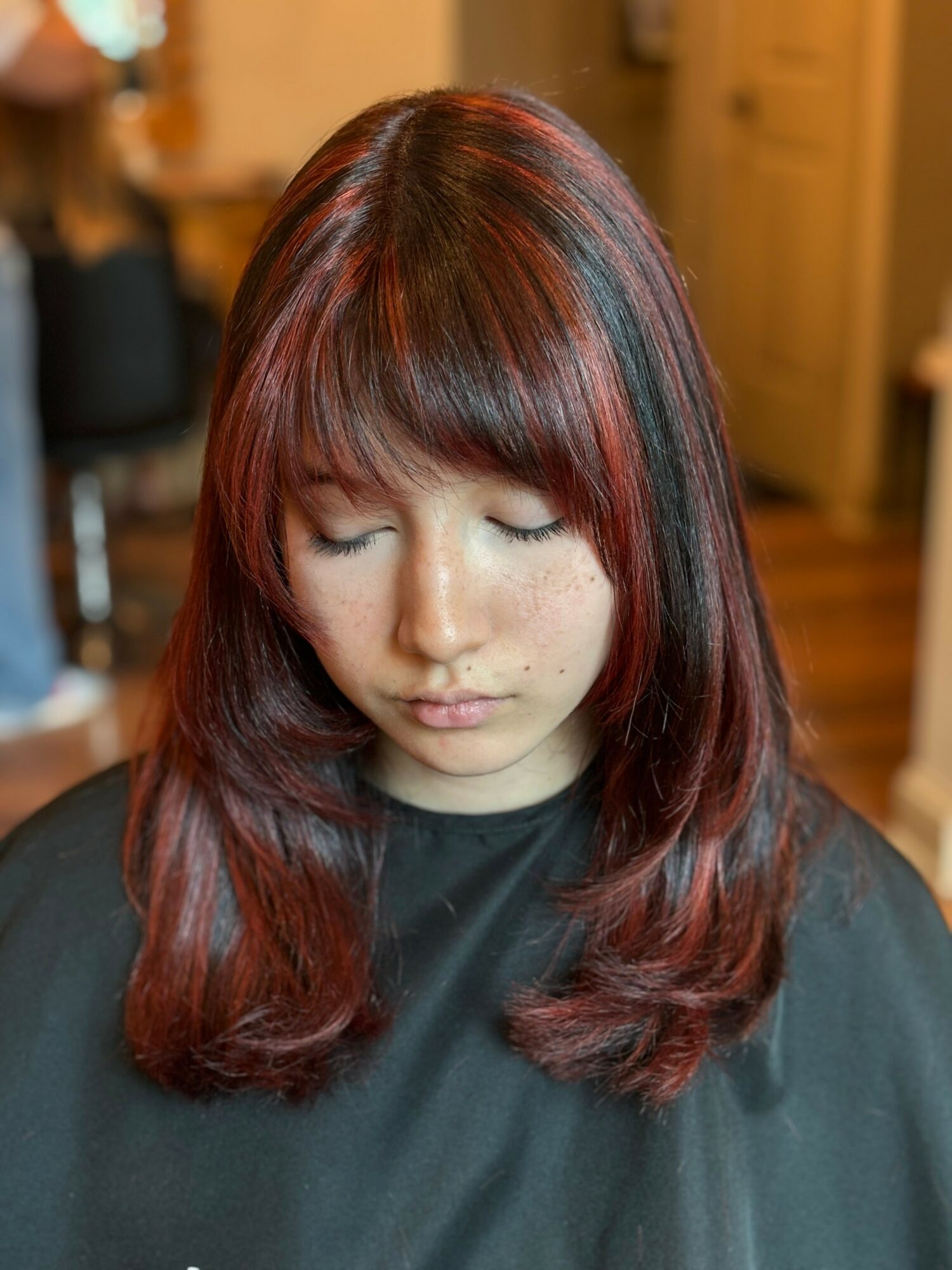 Young woman with shoulder-length red hair and bangs, eyes closed, in a salon setting, wearing a black cape.