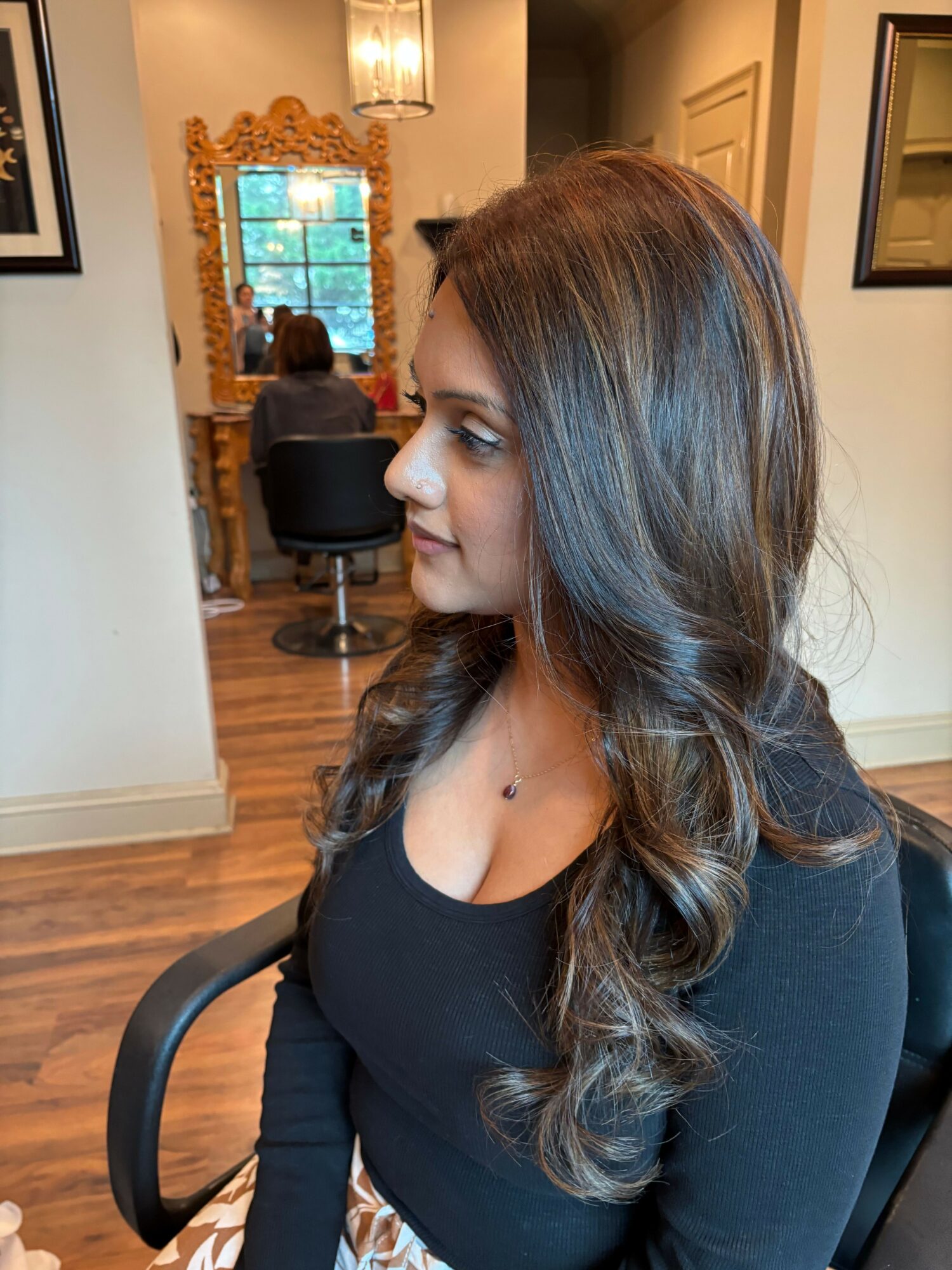 Woman with long, wavy hair looking down, sitting in a room with a mirror and wooden furniture.