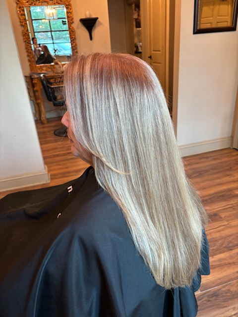 Side view of a woman with long, straight, silver-blonde hair in a salon, wearing a black cape, with a mirror and decor in background.