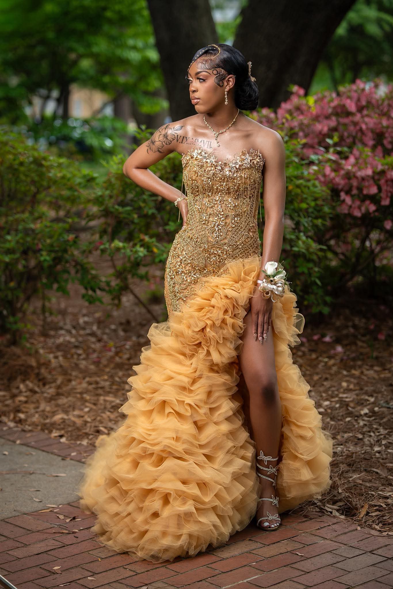 Young woman in a gold, ruffled gown with a fitted, beaded bodice, standing outdoors among trees and pink flowers.