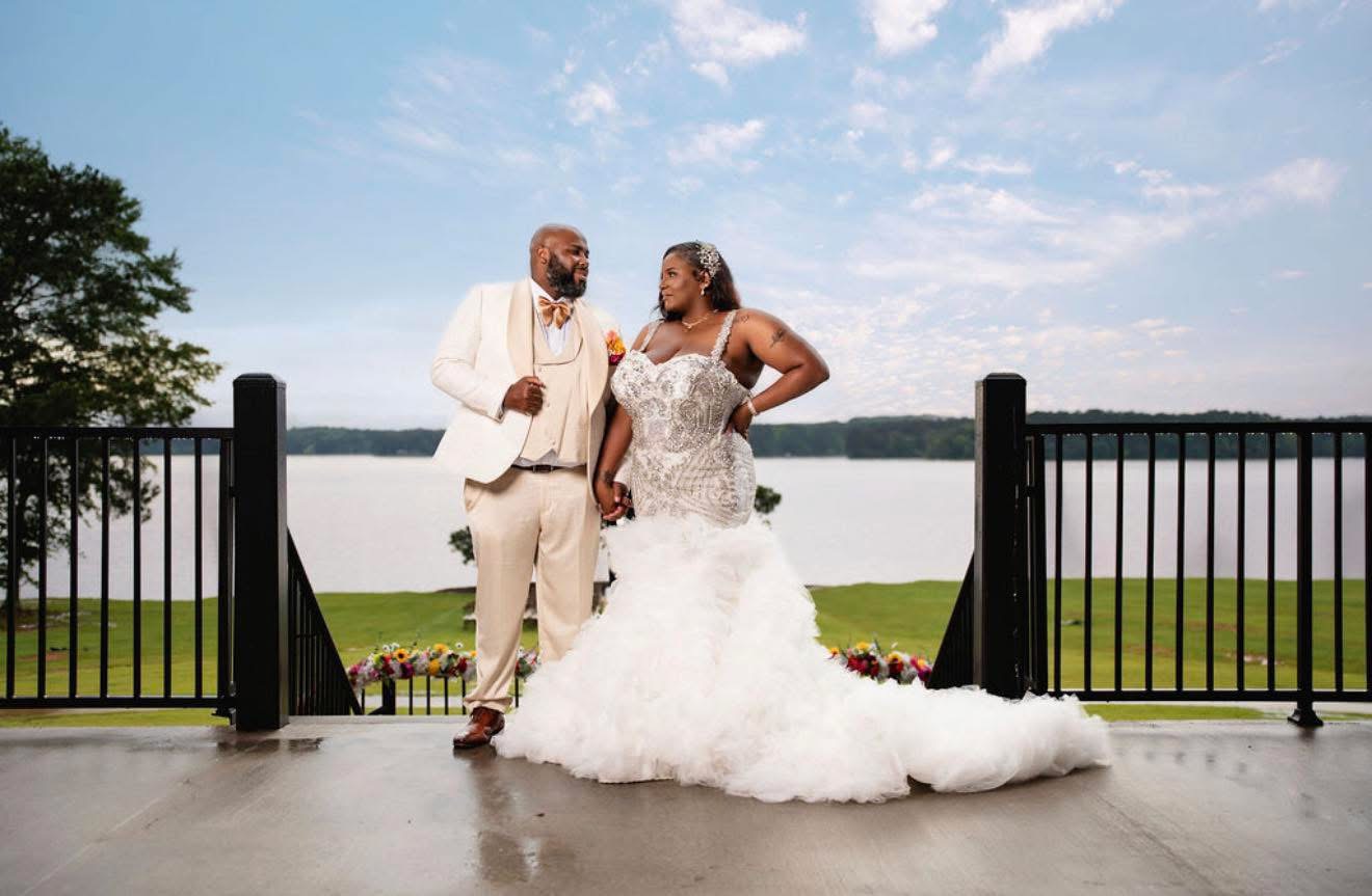 A bride and groom stand outdoors near a lake, facing each other, with a black fence behind them, under a blue sky.