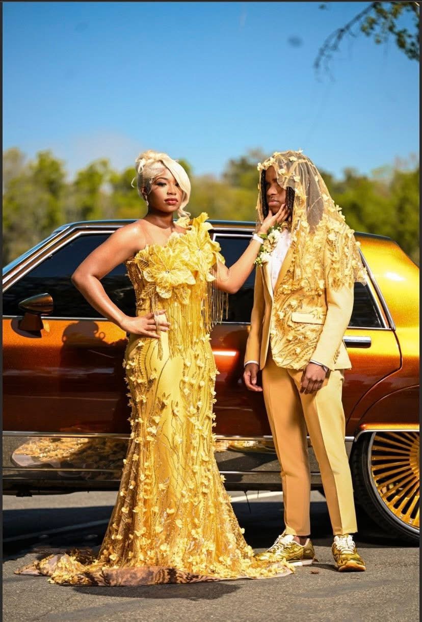 Two women in yellow outfits stand outdoors near a vintage car, one touching the other's face, with trees and blue sky in background.