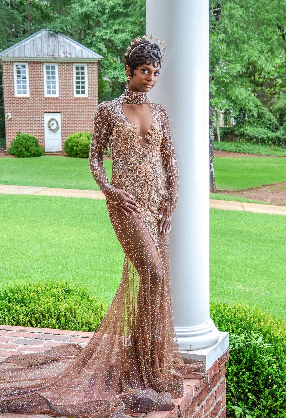 Woman in a long, sheer, embellished gown standing outdoors near a white column, with a brick building and green trees in background.